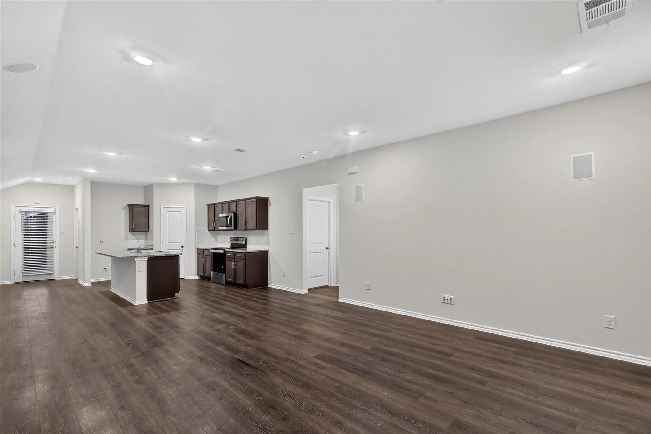 Expansive open-concept living area with wood-finish flooring, recessed lighting, and a kitchen featuring dark wood cabinetry, a center island with a sink, and integrated appliances