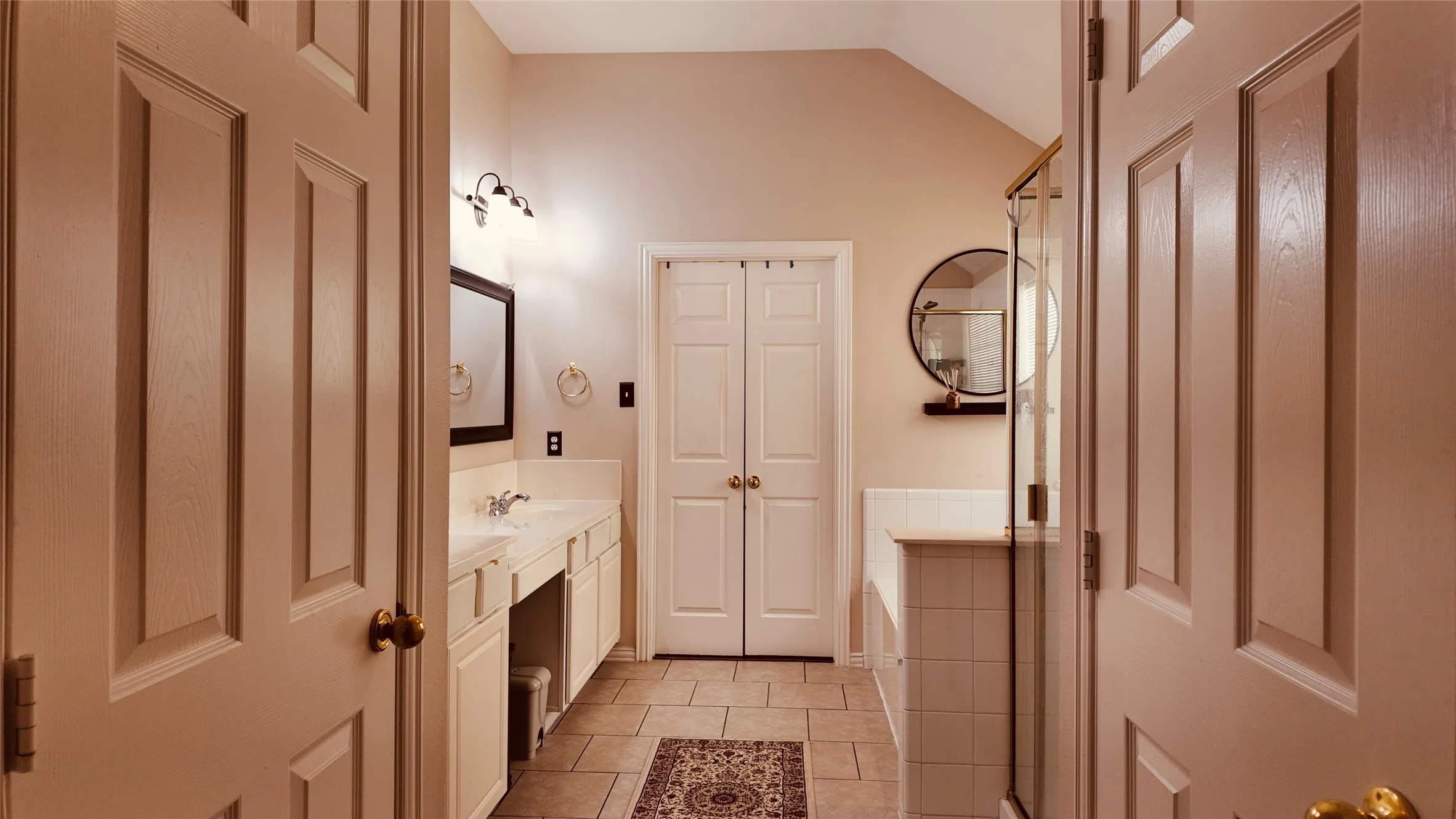 Dual vanity with integrated sinks, tiled flooring, built-in bathtub, glass-enclosed shower, and vaulted ceiling