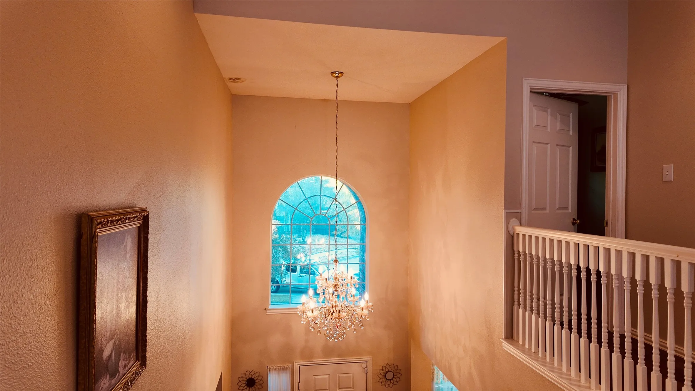 Two-story entryway with a decorative crystal chandelier, a large arched window, and a white wood-spindle railing