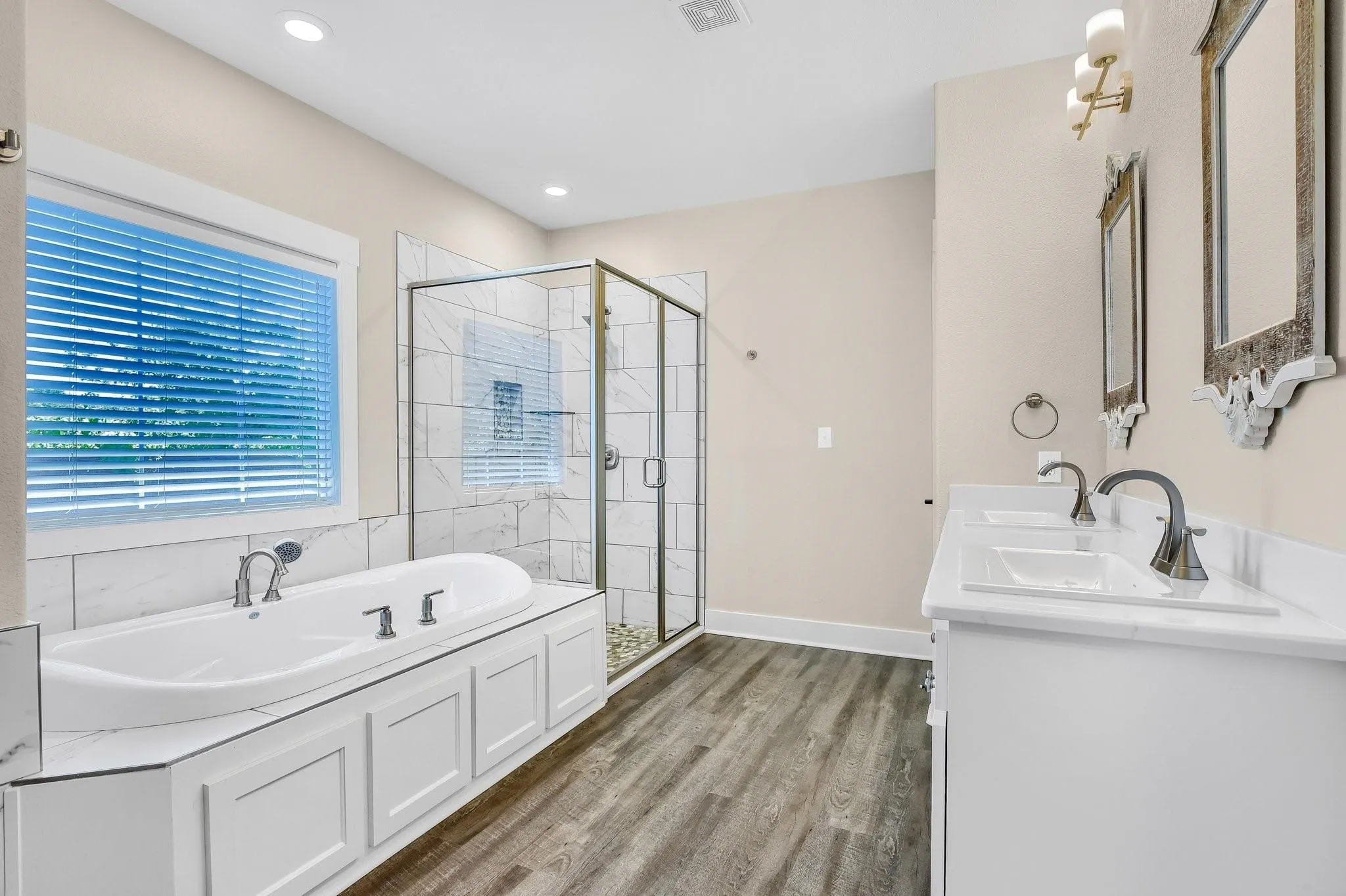 Soaking tub with white paneled surround, walk-in shower with clear glass enclosure and tile surround, dual vanity with white countertops, wood-finish flooring, and recessed lighting