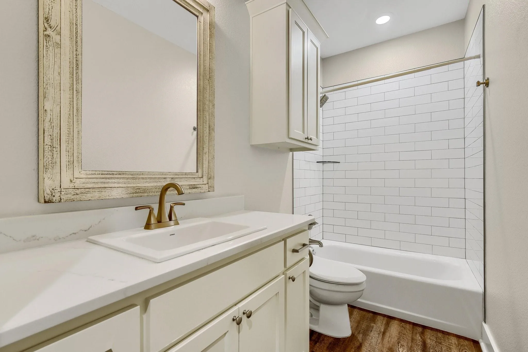 White subway tile shower surround, single vanity with white countertop, brass-finish faucet, recessed lighting, and wood-finish flooring