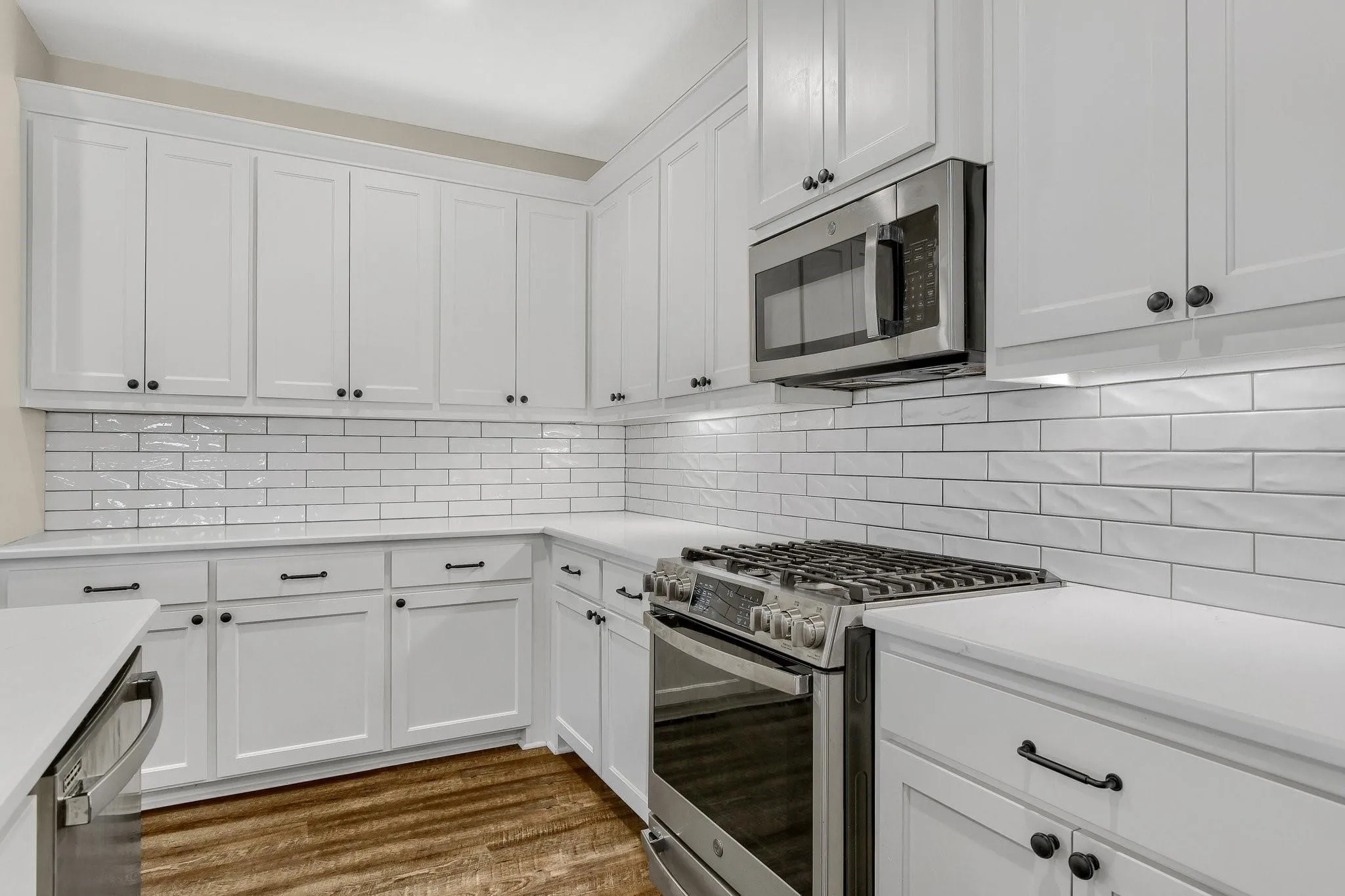 White cabinetry with black hardware, subway tile backsplash, stainless steel gas range, stainless steel microwave, and wood-finish flooring