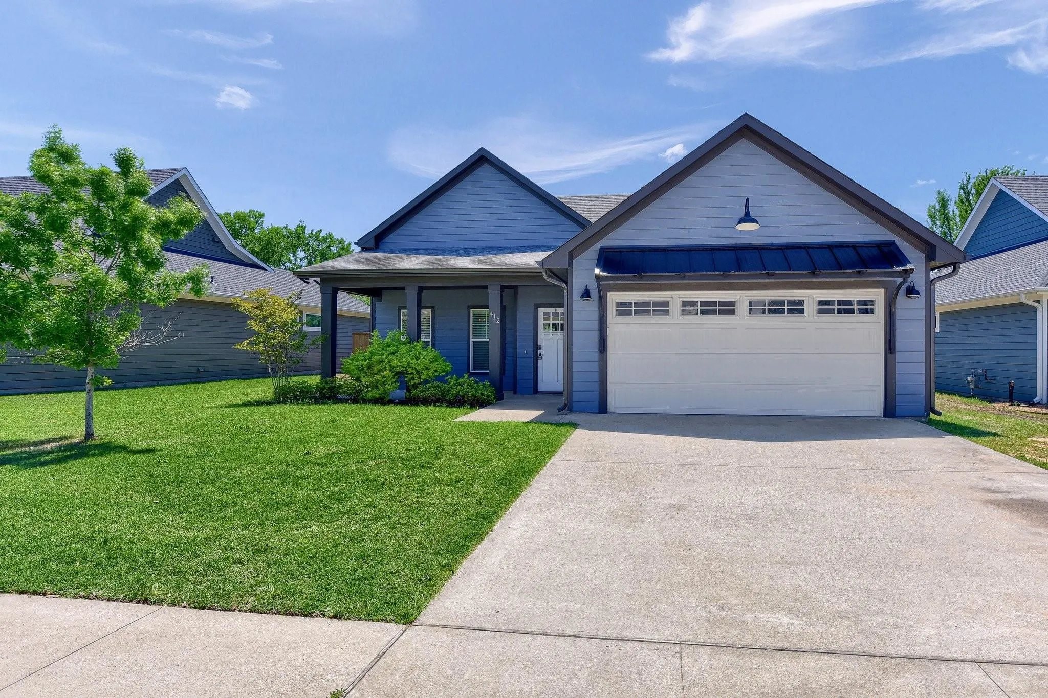 Light blue siding with dark trim accents, covered front porch, attached two-car garage, concrete driveway, and a front yard with manicured lawn