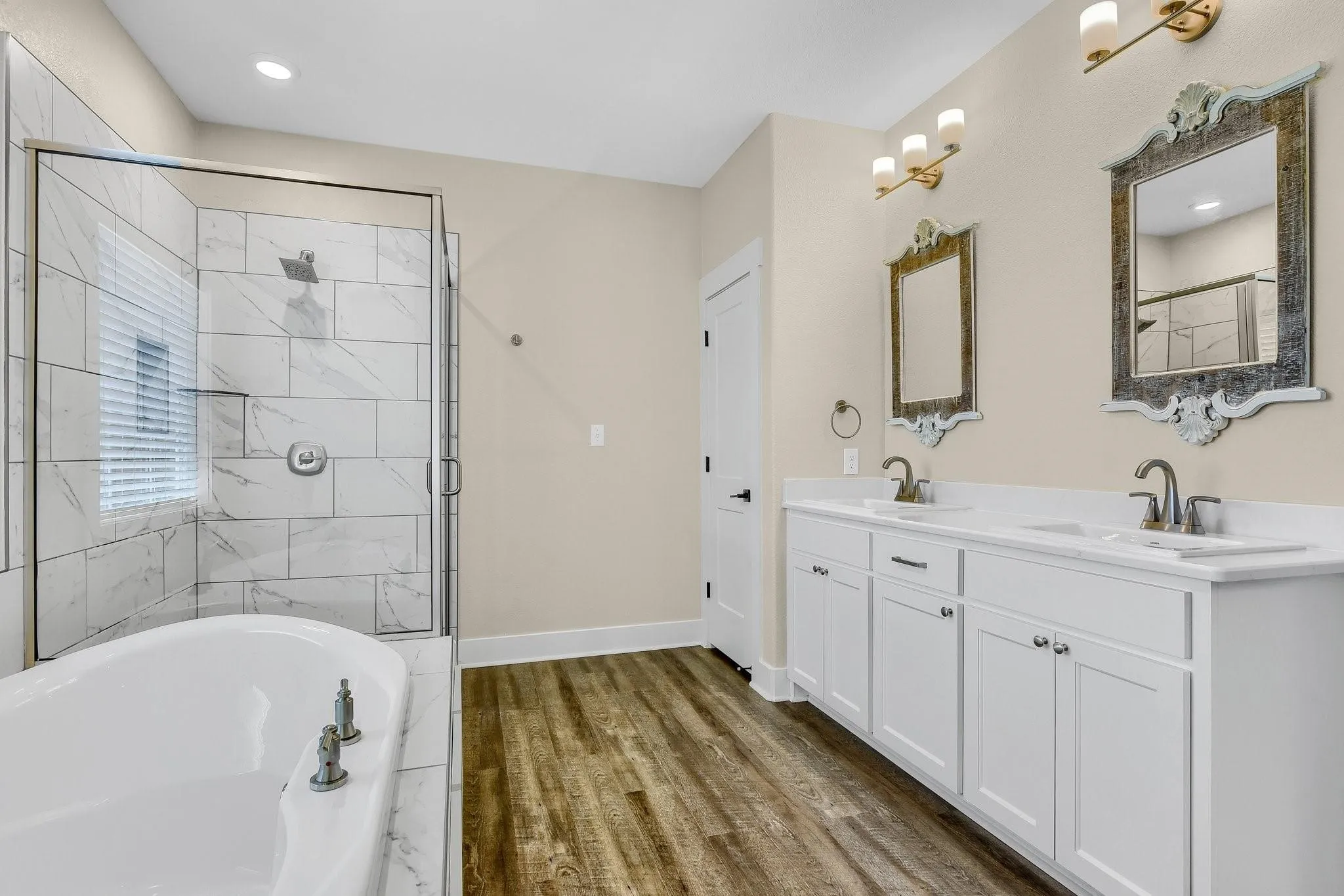 Dual vanity with white cabinetry and integrated sinks, a walk-in shower with tile surround and glass enclosure, a freestanding soaking tub, and wood-finish flooring