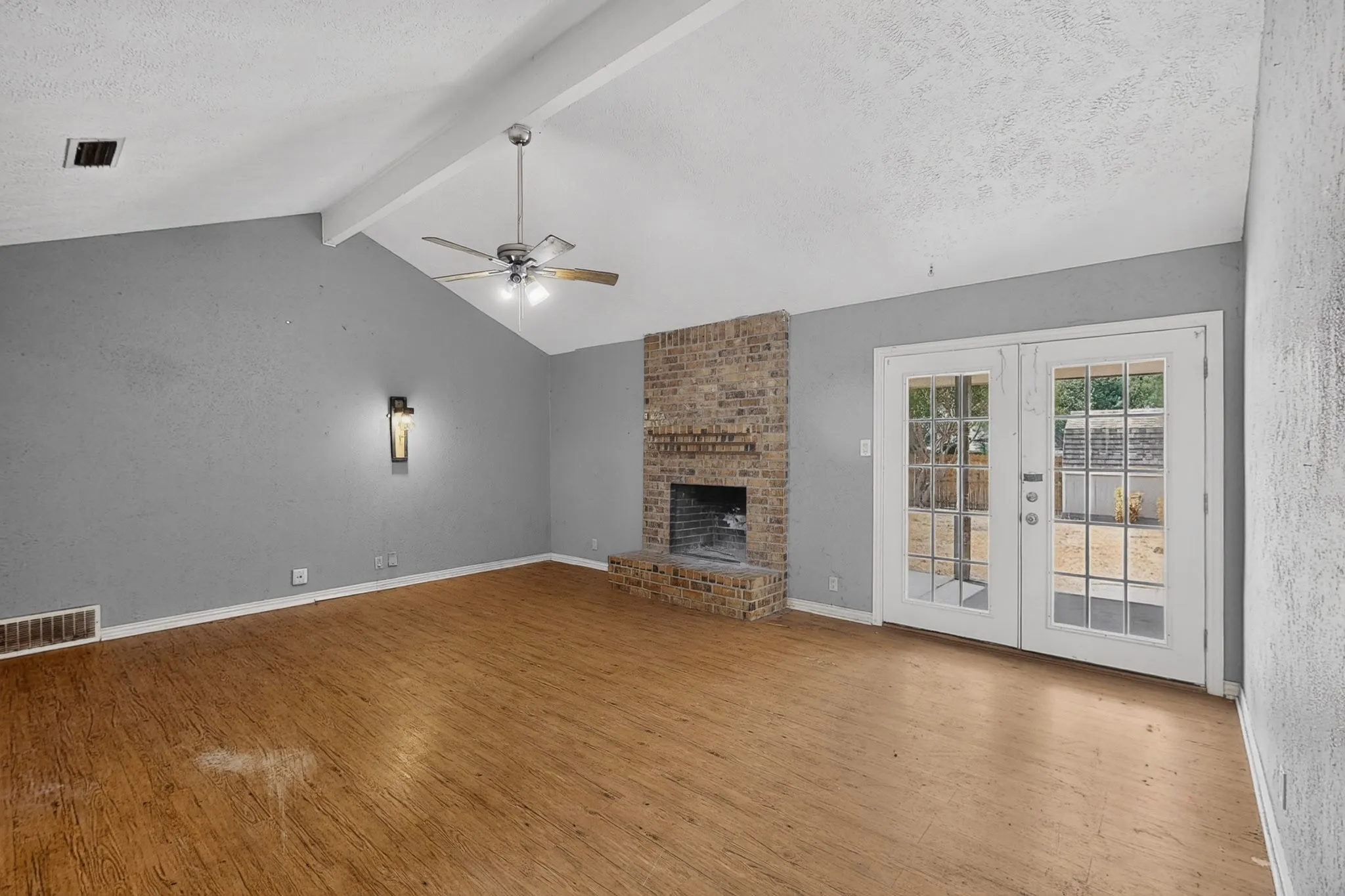 Vaulted ceiling with exposed beams, brick fireplace, wood-finish flooring, ceiling fan, and double French doors with divided lites