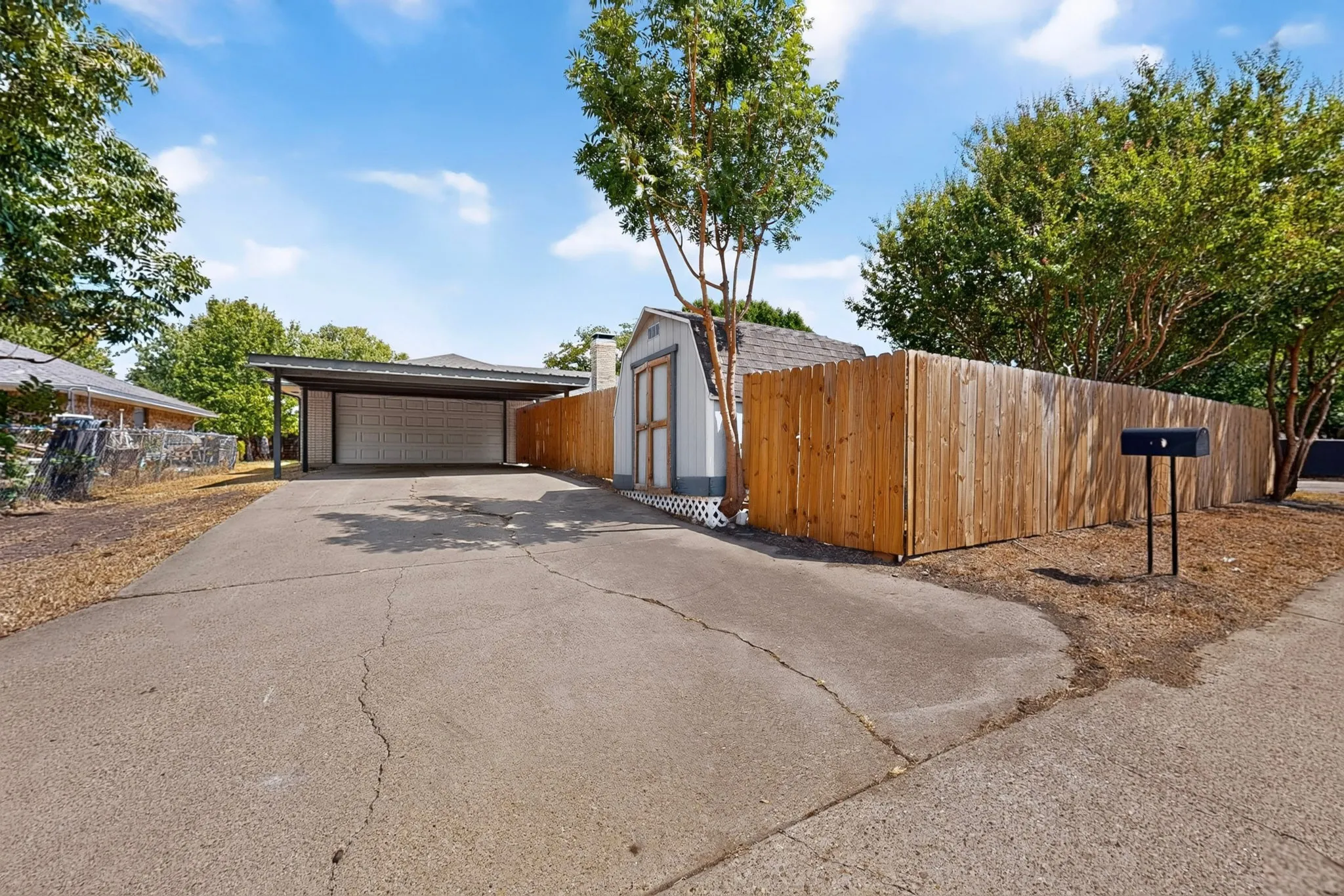 Paved driveway leading to a covered carport, wood privacy fence, and an exterior storage shed