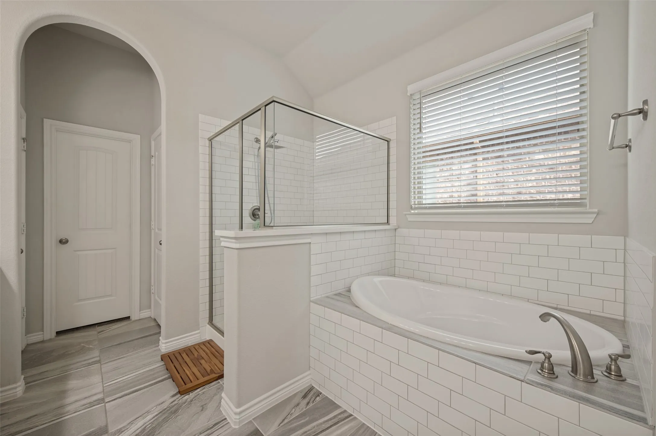 Freestanding soaking tub with subway tile surround, separate glass-enclosed shower, tiled flooring, and arched doorway
