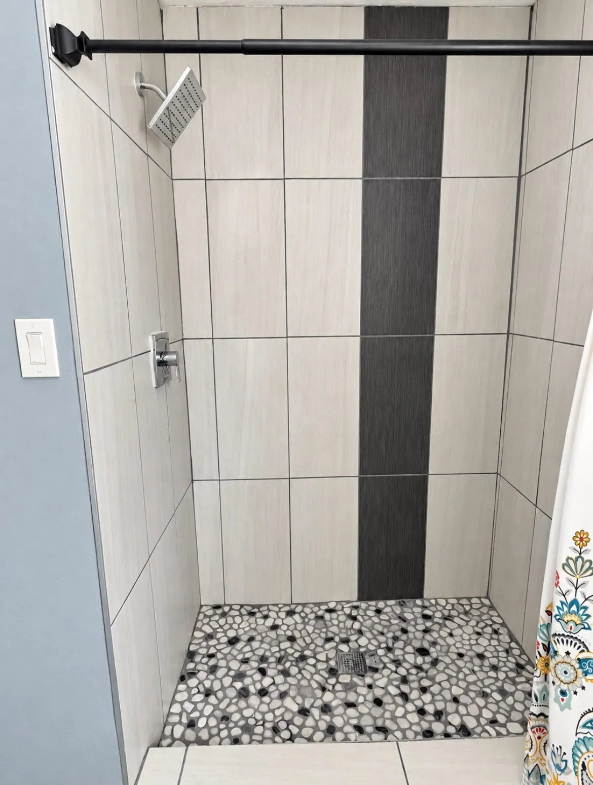 Shower with light-toned rectangular wall tiles, a contrasting dark accent tile column, and a pebble mosaic floor