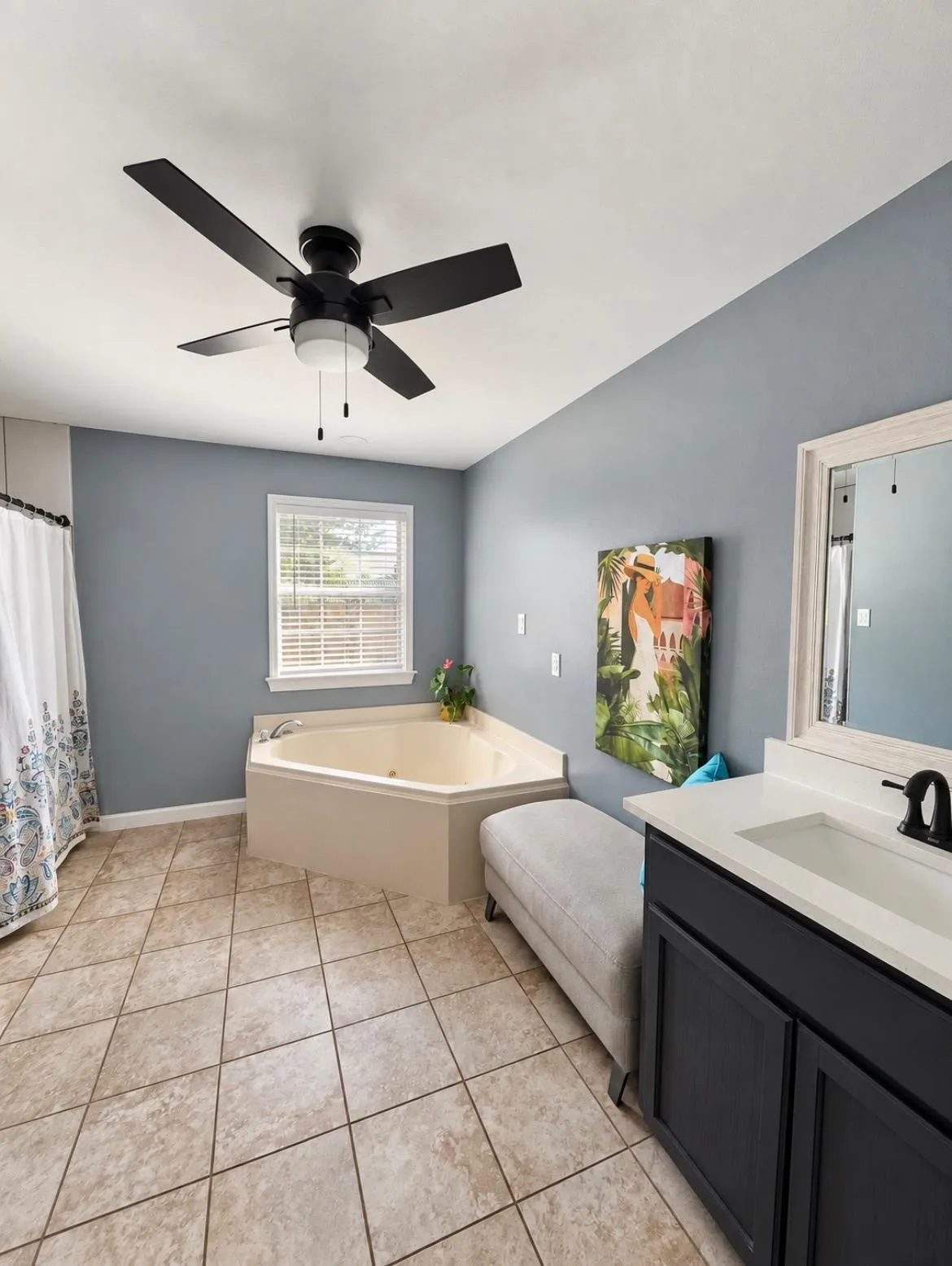 Corner soaking tub, tiled flooring, single vanity with integrated sink, black ceiling fan with lighting, and window with blinds