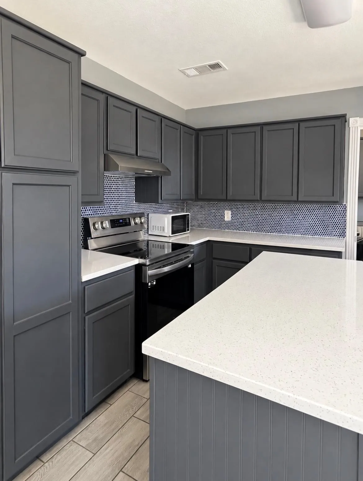 Kitchen featuring dark cabinetry, light-toned countertops, a stainless steel range with overhead vent, and a blue mosaic tile backsplash