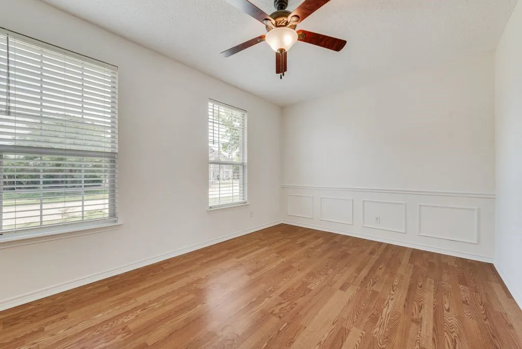 Spacious room featuring wood-finish flooring, white walls with wainscoting, two windows with blinds, and a ceiling fan with integrated lighting
