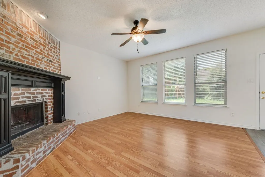 Living area featuring a brick-face fireplace with a dark wood mantel, wood-finish flooring, three windows with blinds, and a ceiling fan