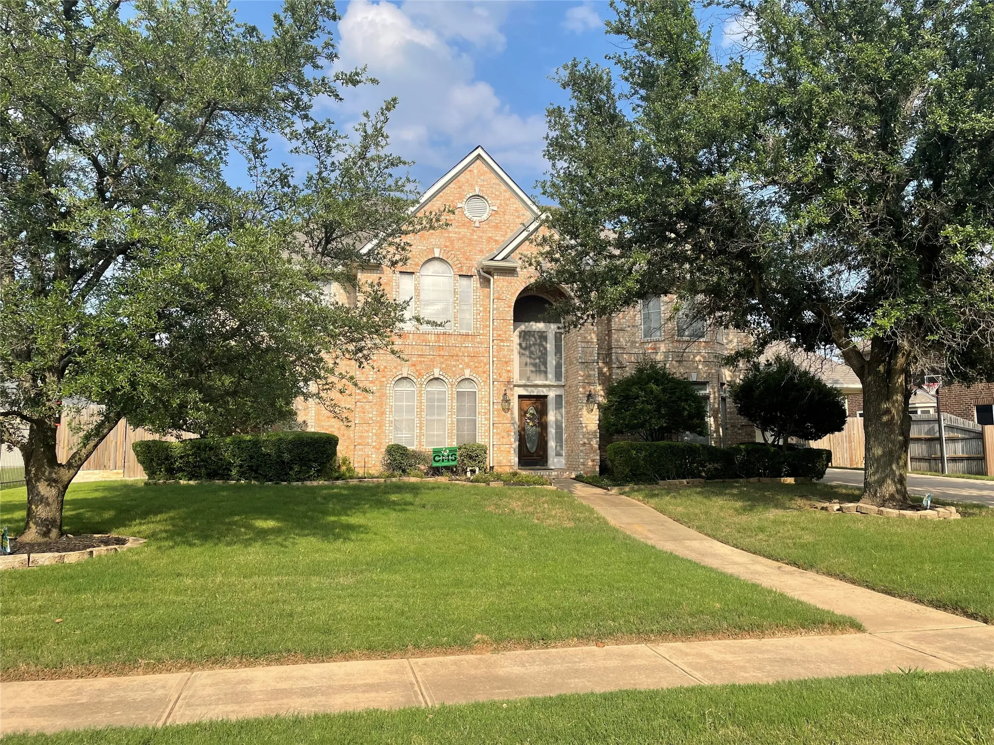 Brick facade residence with an arched entry, mature trees, a manicured lawn, and a concrete pathway