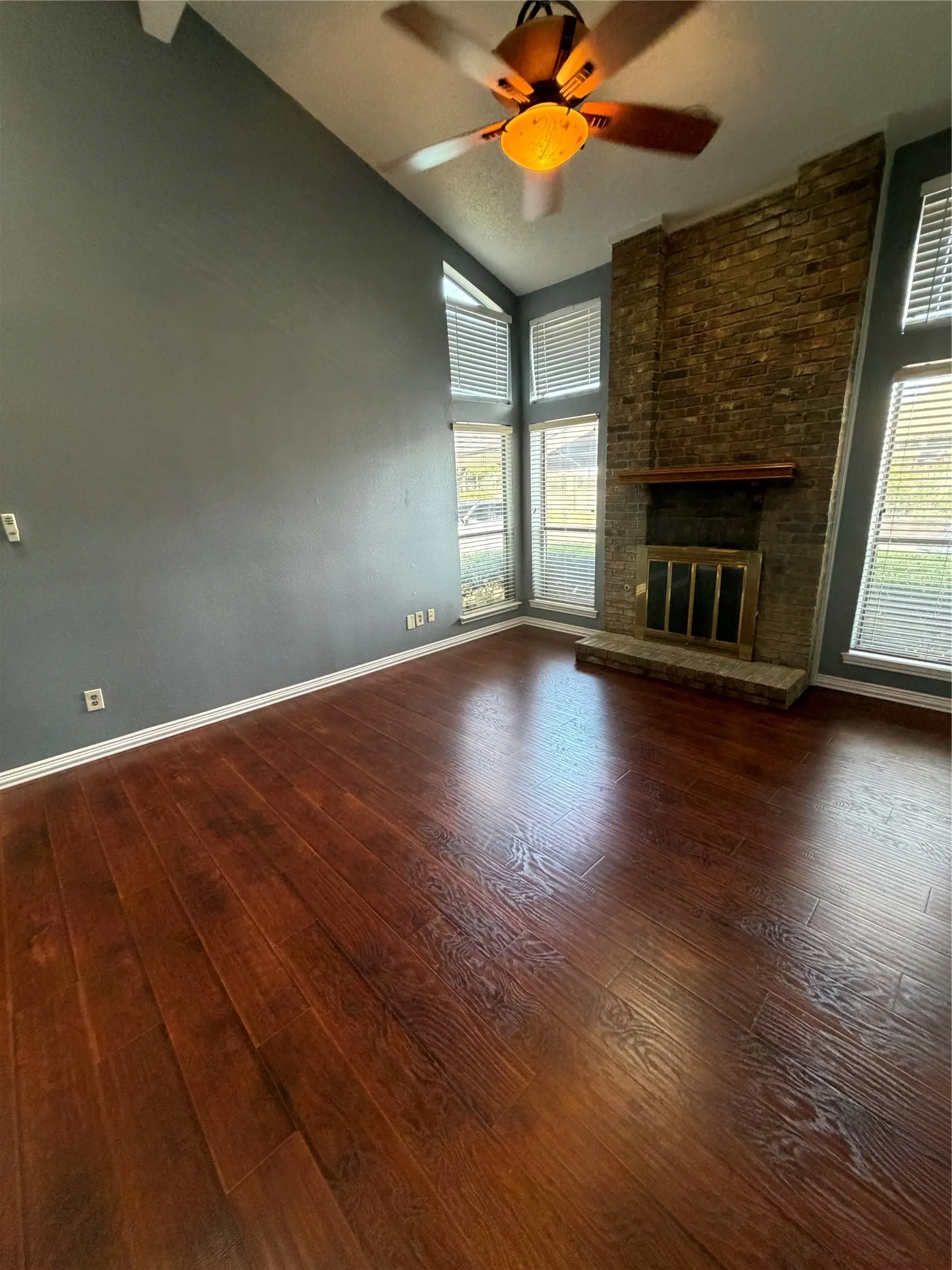 Vaulted ceiling, wood-finish flooring, brick fireplace with mantel, ceiling fan with light fixture, and multiple windows with blinds
