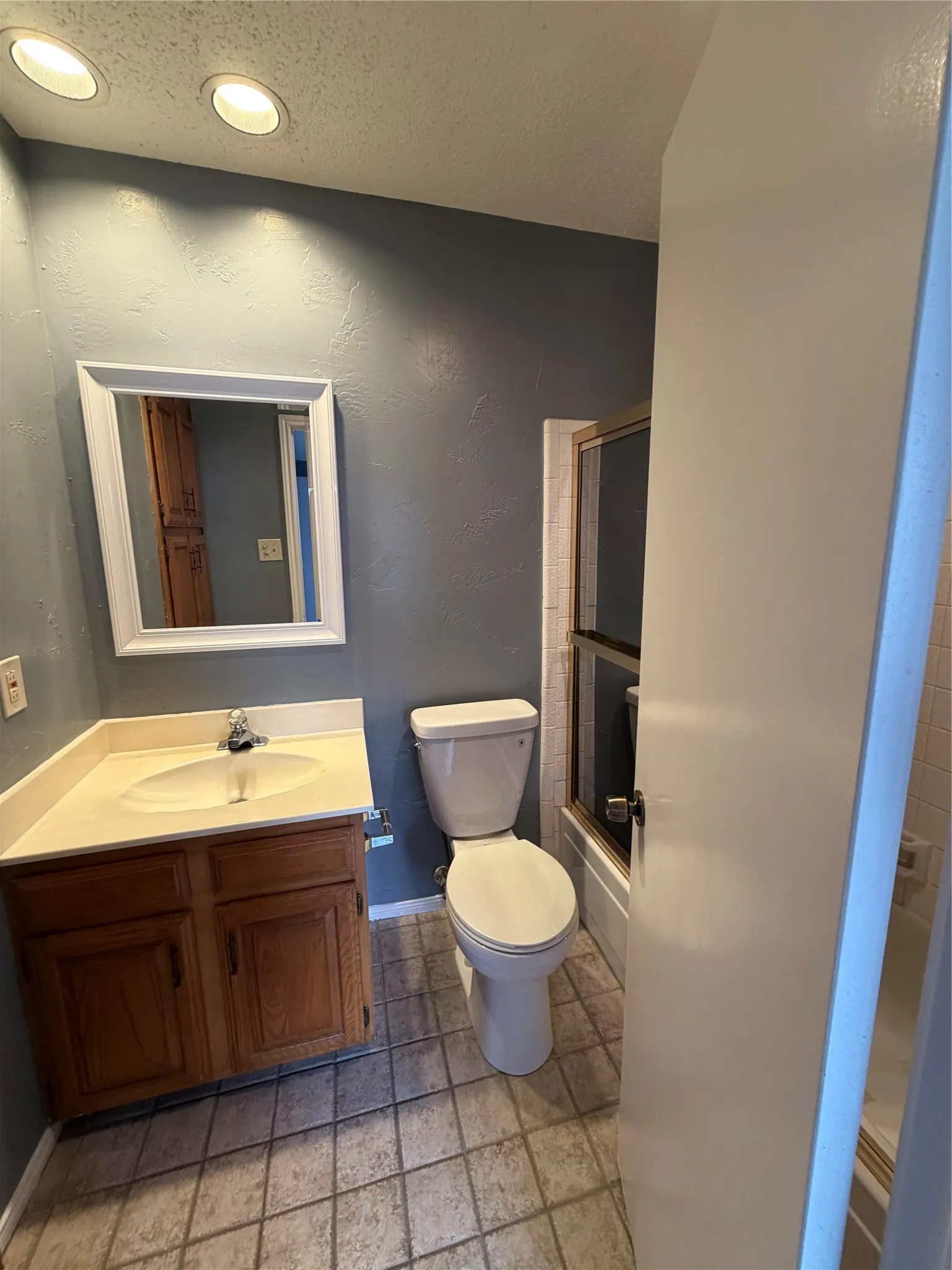 Recessed ceiling lighting, wood-finish vanity with countertop, white framed mirror, toilet, and tiled flooring