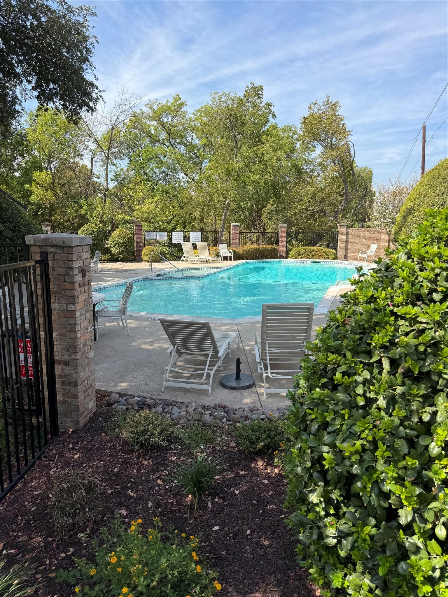 Swimming pool with concrete surround, surrounded by mature trees and shrubs, featuring a black metal fence with brick pillars