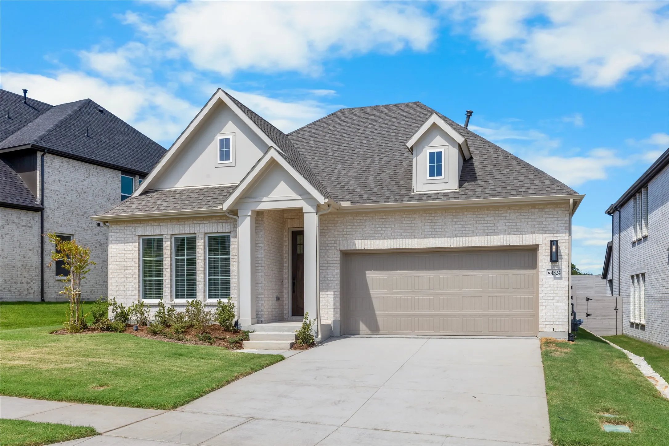 White brick exterior with a gray shingle roof, featuring a dormer window and an attached two-car garage