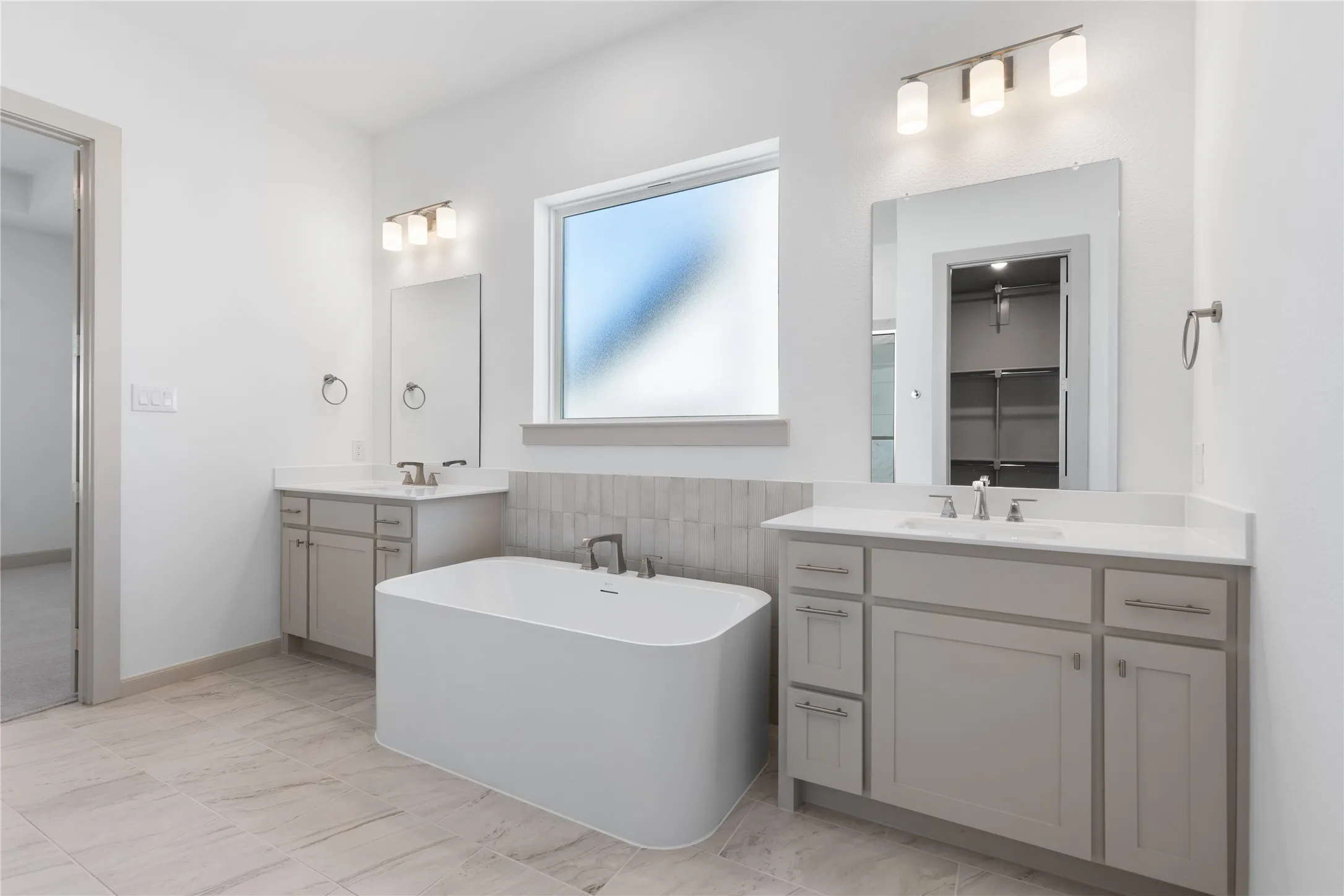 Freestanding soaking tub with a tiled surround, dual vanities with integrated sinks, and neutral-toned tile flooring