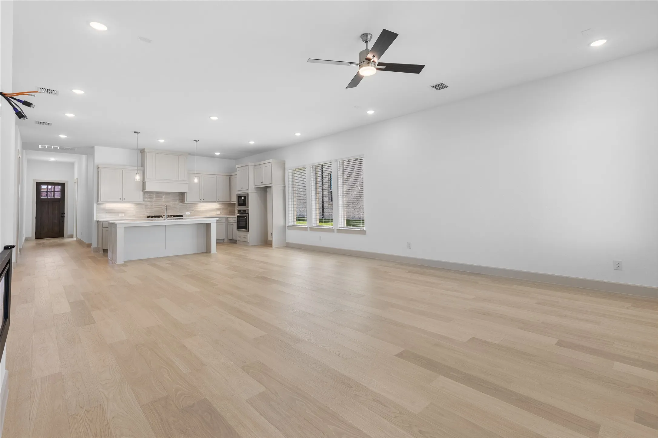 Expansive open-concept space featuring light wood-finish flooring, a kitchen island with pendant lighting, recessed ceiling lights, and contemporary ceiling fan