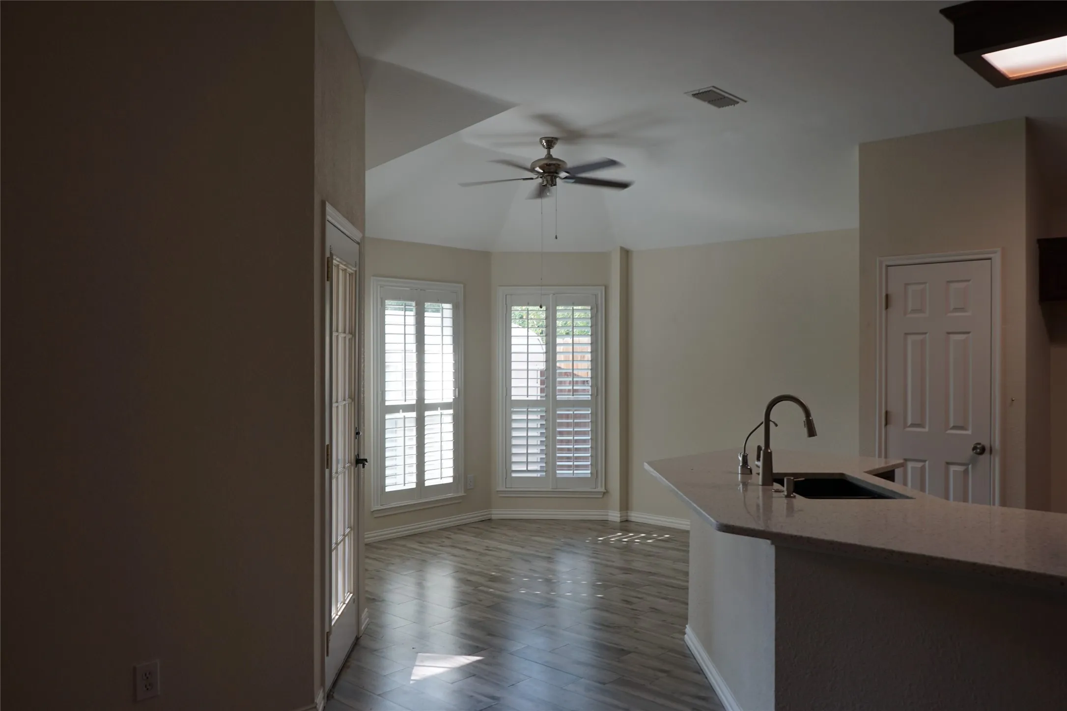 Bay window with plantation shutters, wood-finish flooring, vaulted ceiling, built-in ceiling fan, and a kitchen island with sink and faucet