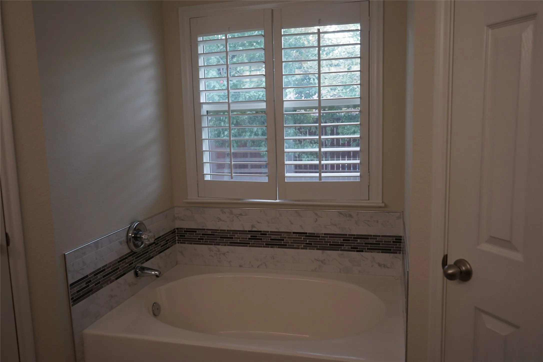 Built-in soaking tub featuring a marble-look surround with a glass mosaic accent, and a window with white plantation shutters