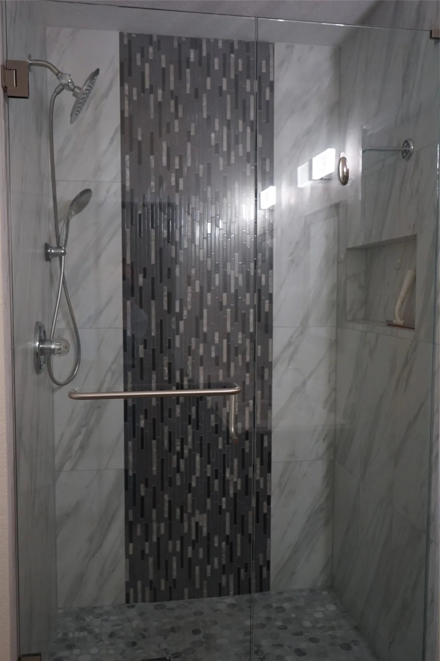 Glass-enclosed shower featuring a tiled accent wall, dual shower heads, a built-in niche, and pebble-finish flooring