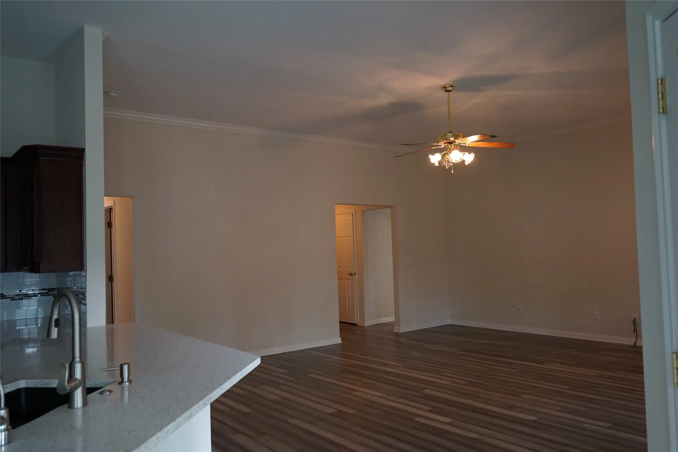 Wood-finish flooring, crown molding, ceiling fan with light fixture, kitchen with dark wood-finish cabinetry, and light-toned backsplash
