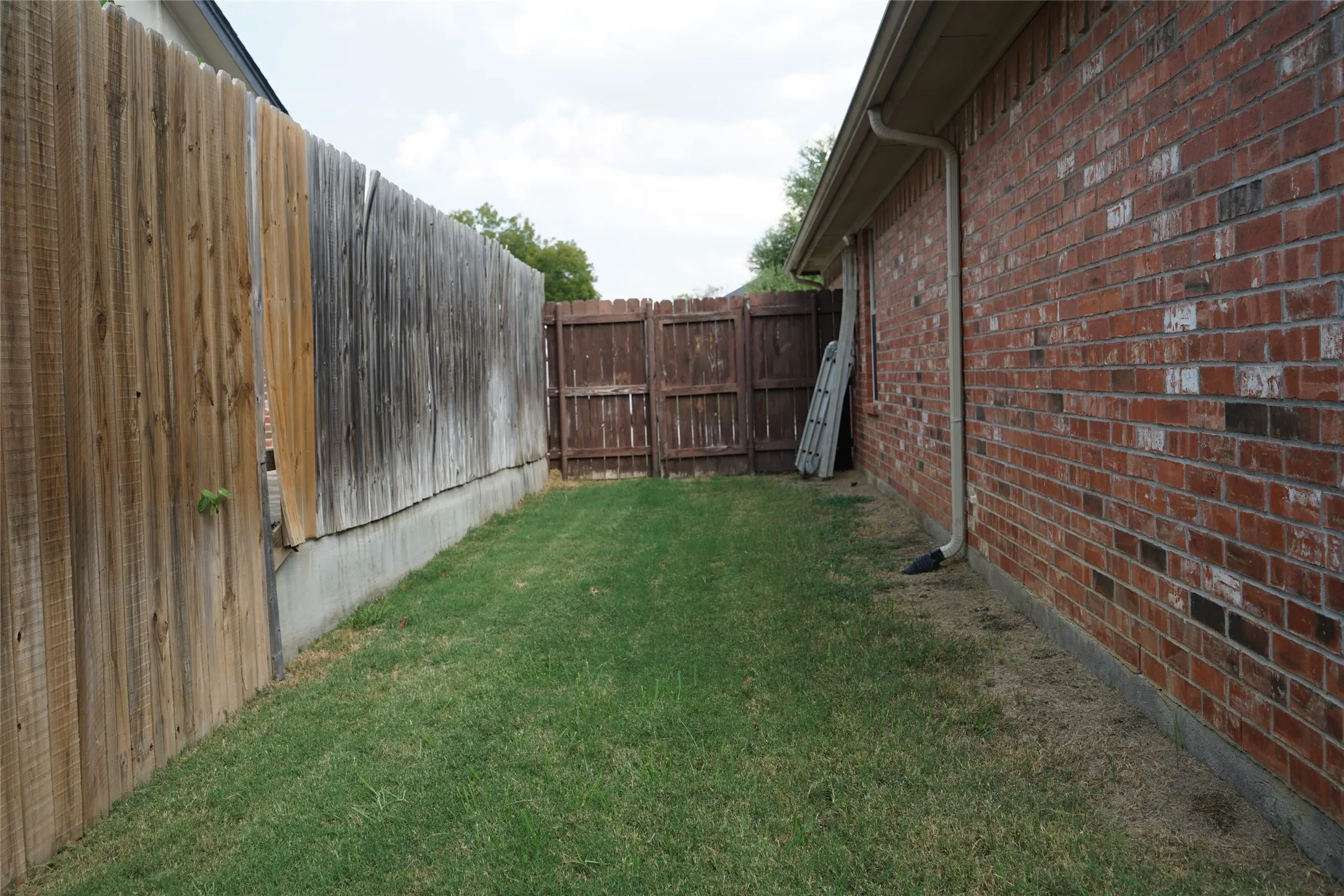 Brick exterior wall with a rain gutter system, bordered by a wood privacy fence on a concrete base