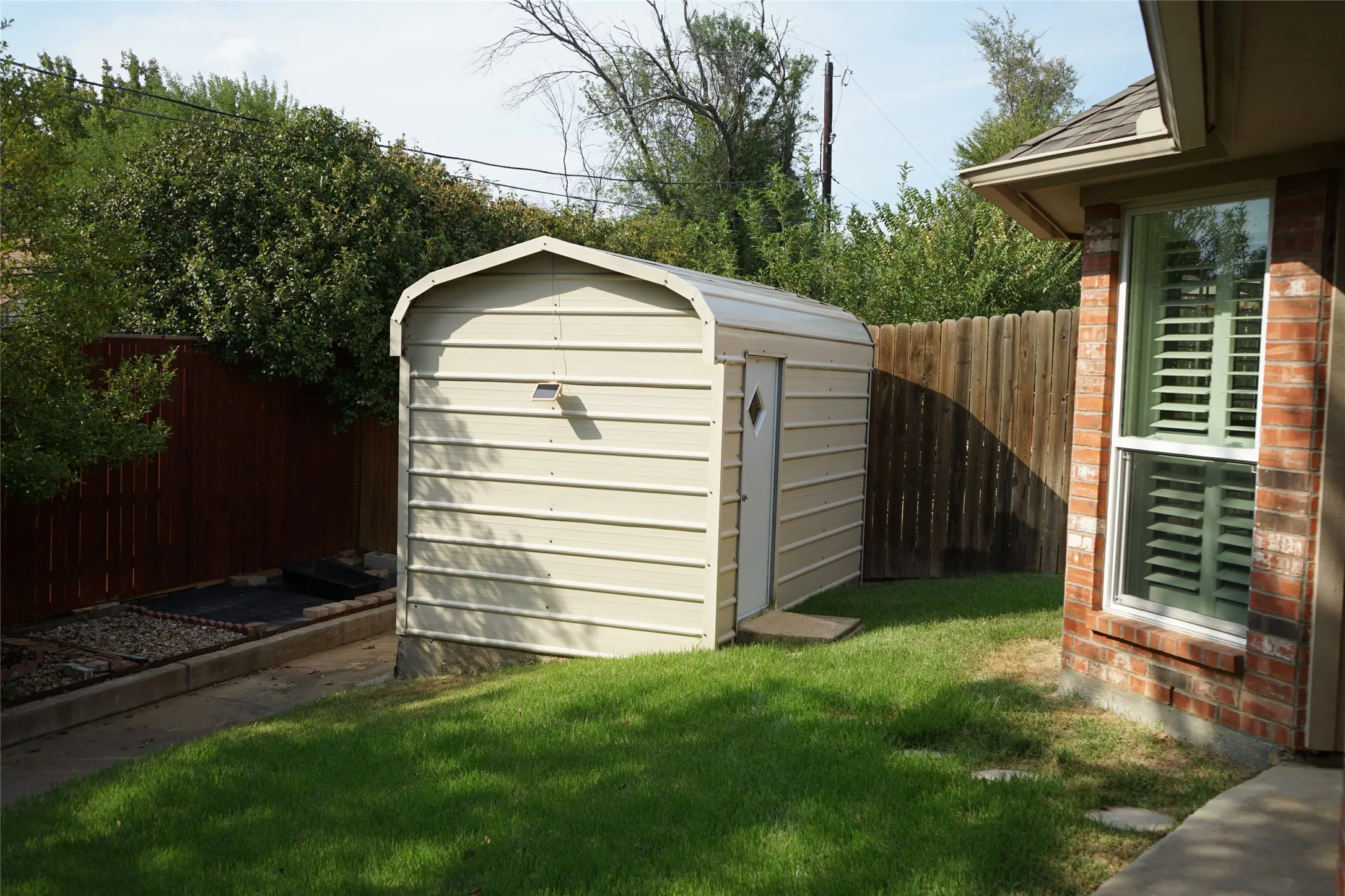 Gabled storage shed, wood privacy fencing, brick exterior, single-hung window with shutters, and manicured lawn