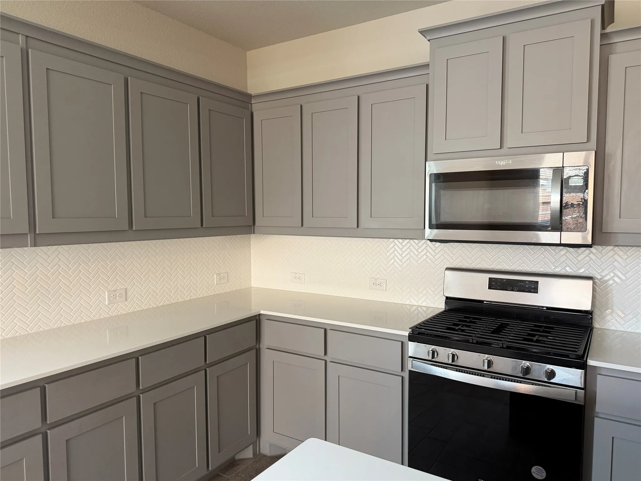 Neutral-toned cabinetry, white herringbone tile backsplash, stainless steel microwave, gas range, and light-colored countertops
