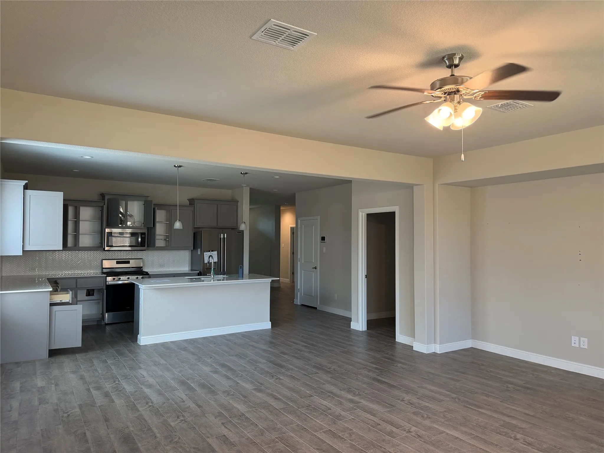 Open-concept living area featuring wood-finish flooring, a ceiling fan with integrated lighting, and a kitchen with stainless steel appliances, grey cabinetry, and an island with a sink