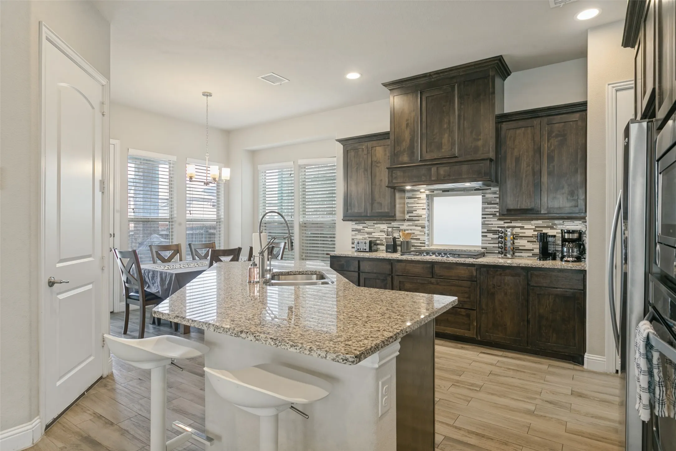 Kitchen featuring an island with a built-in sink, wood-finish flooring, dark wood cabinetry, a tile backsplash, and recessed lighting