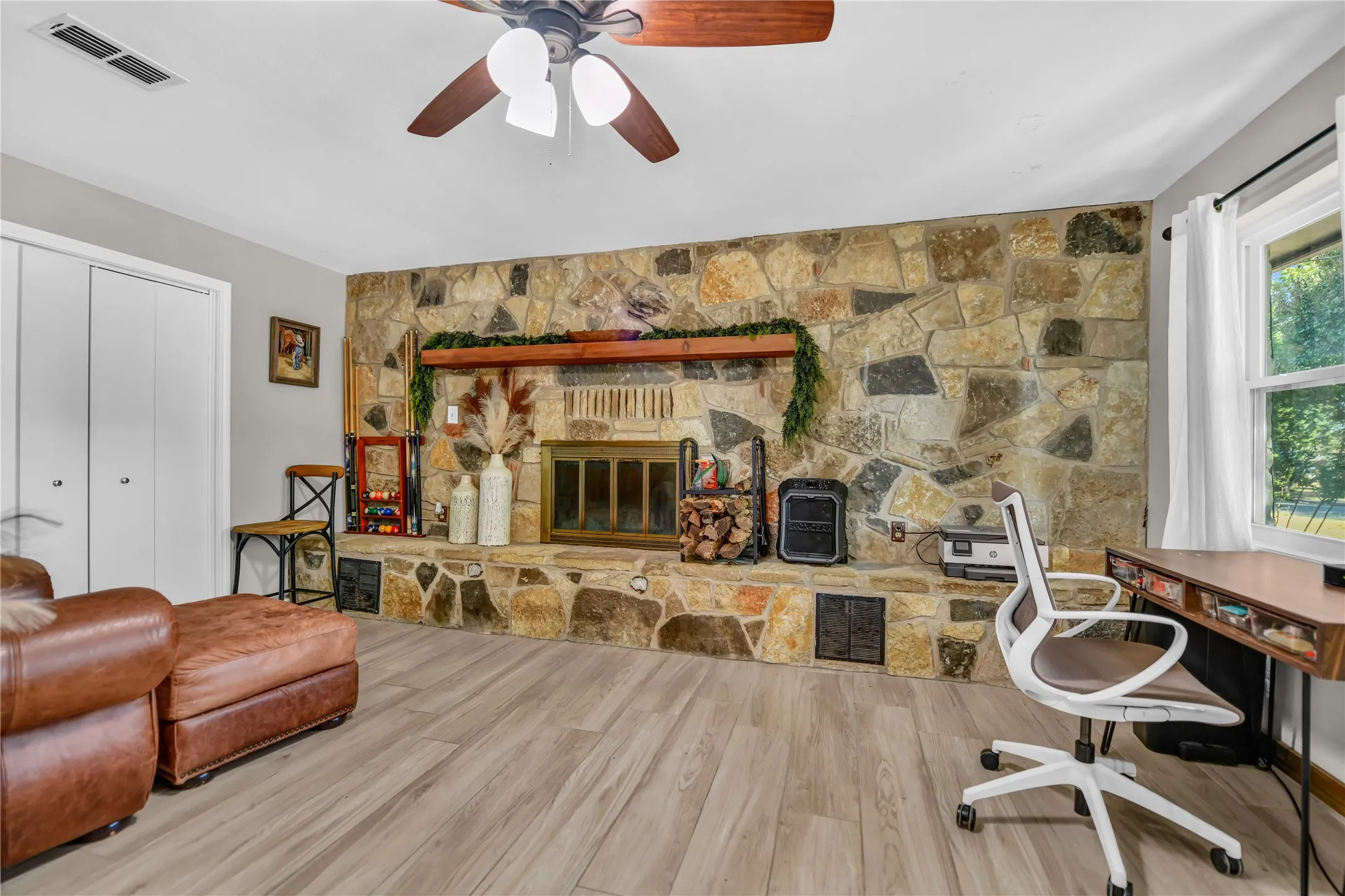 Full-height stone-clad fireplace with wood mantle, wood-finish flooring, ceiling fan, and window with natural light
