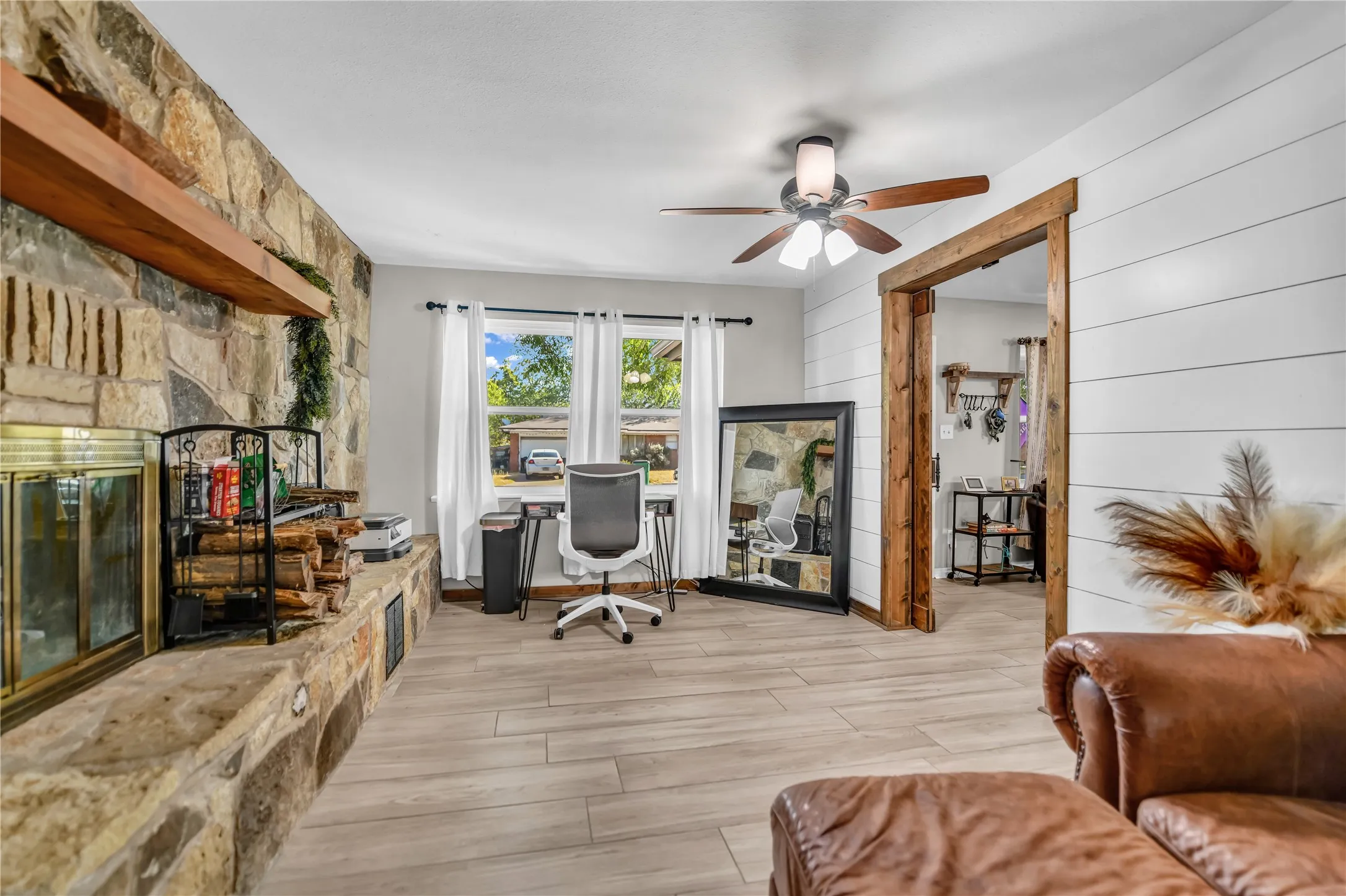 Stone-clad fireplace with a wood mantel, wood-finish flooring, white shiplap accent wall, and a ceiling fan with light fixture