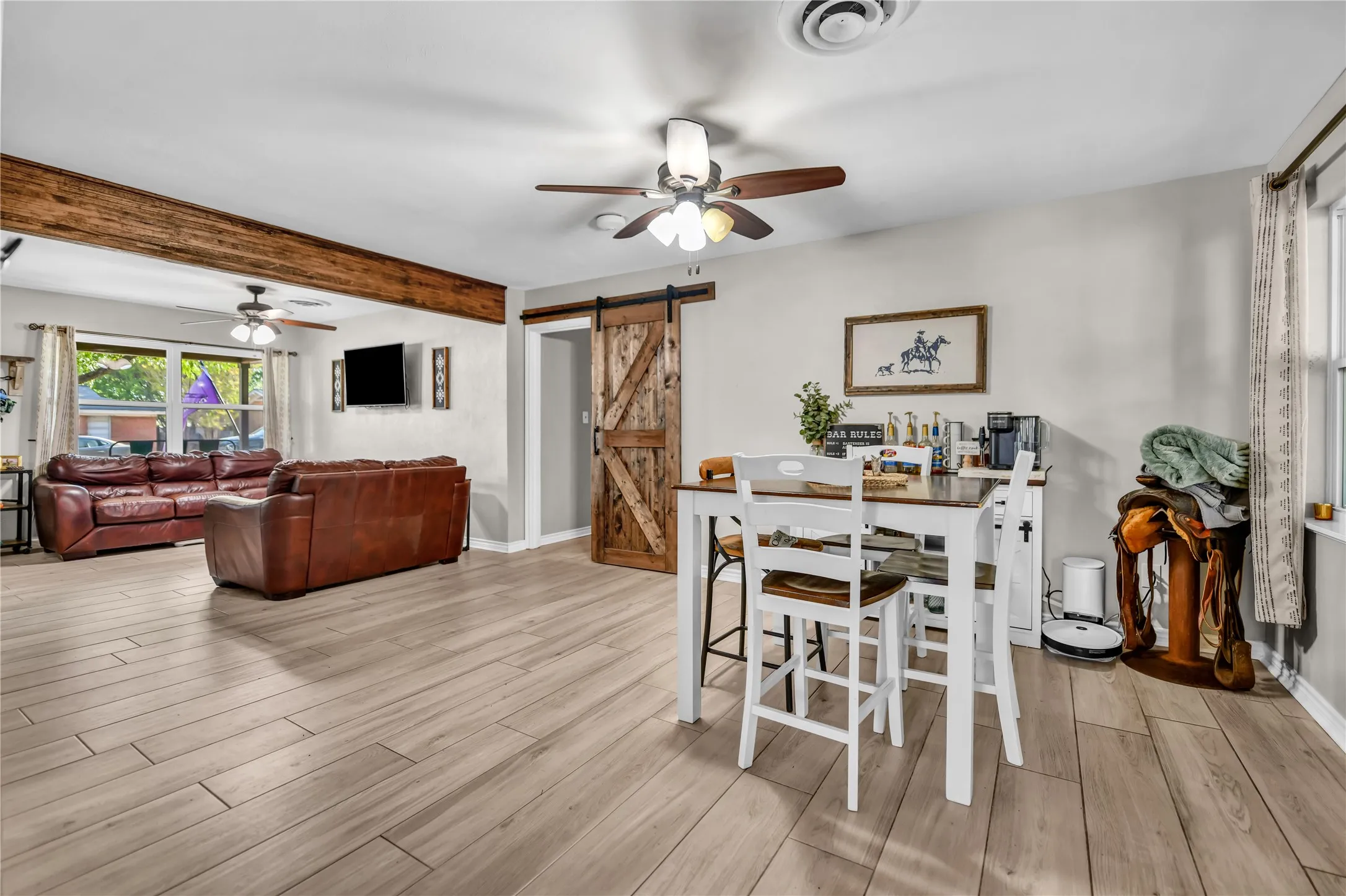 Light wood-finish flooring throughout, ceiling fan with light fixture, exposed wood beam, large window with curtains, and a wood-finish barn door