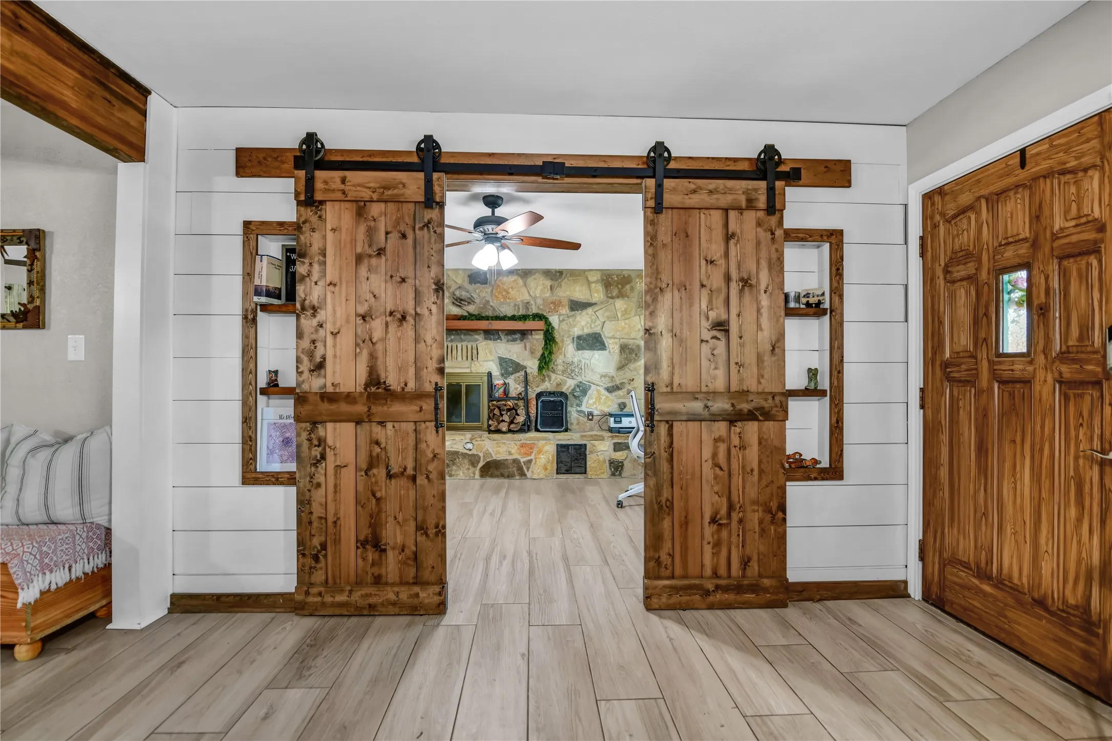 Wood-finish plank flooring, custom wood barn doors, shiplap wall accent, stone-clad fireplace, and a wood ceiling fan
