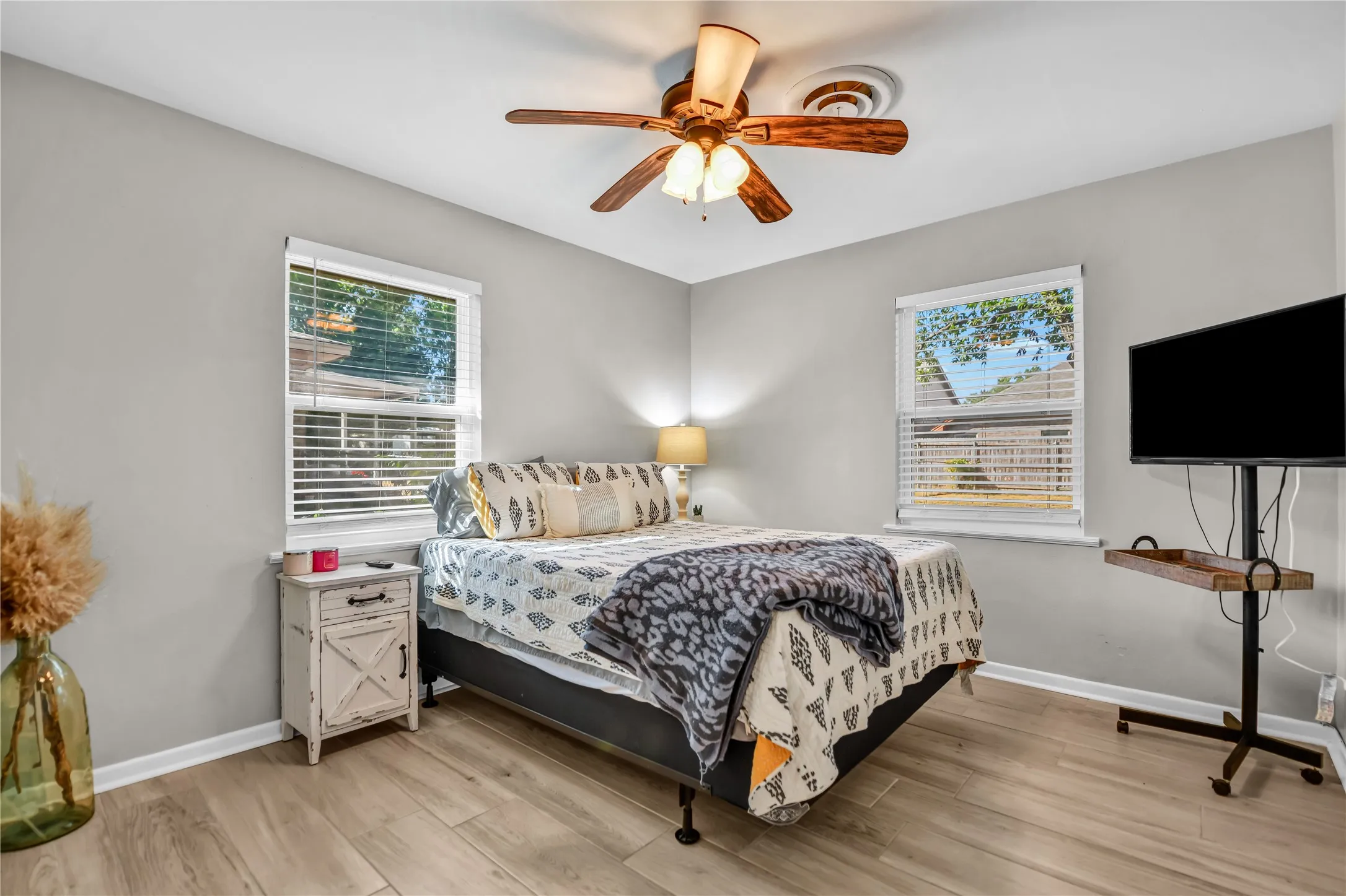Wood-finish flooring, light gray wall paint, two windows with blinds, and a ceiling fan with integrated lighting