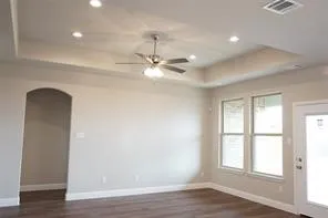 Tray ceiling with recessed lighting, ceiling fan, wood-finish flooring, multiple windows, and an arched doorway