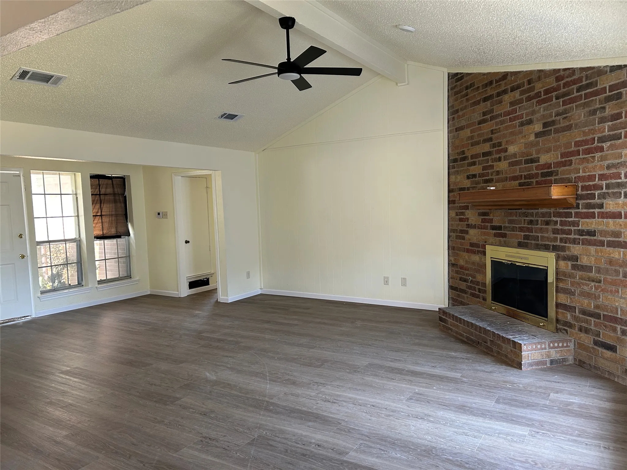 Spacious living area featuring wood-finish flooring, a vaulted ceiling, and a brick fireplace with a wood mantle
