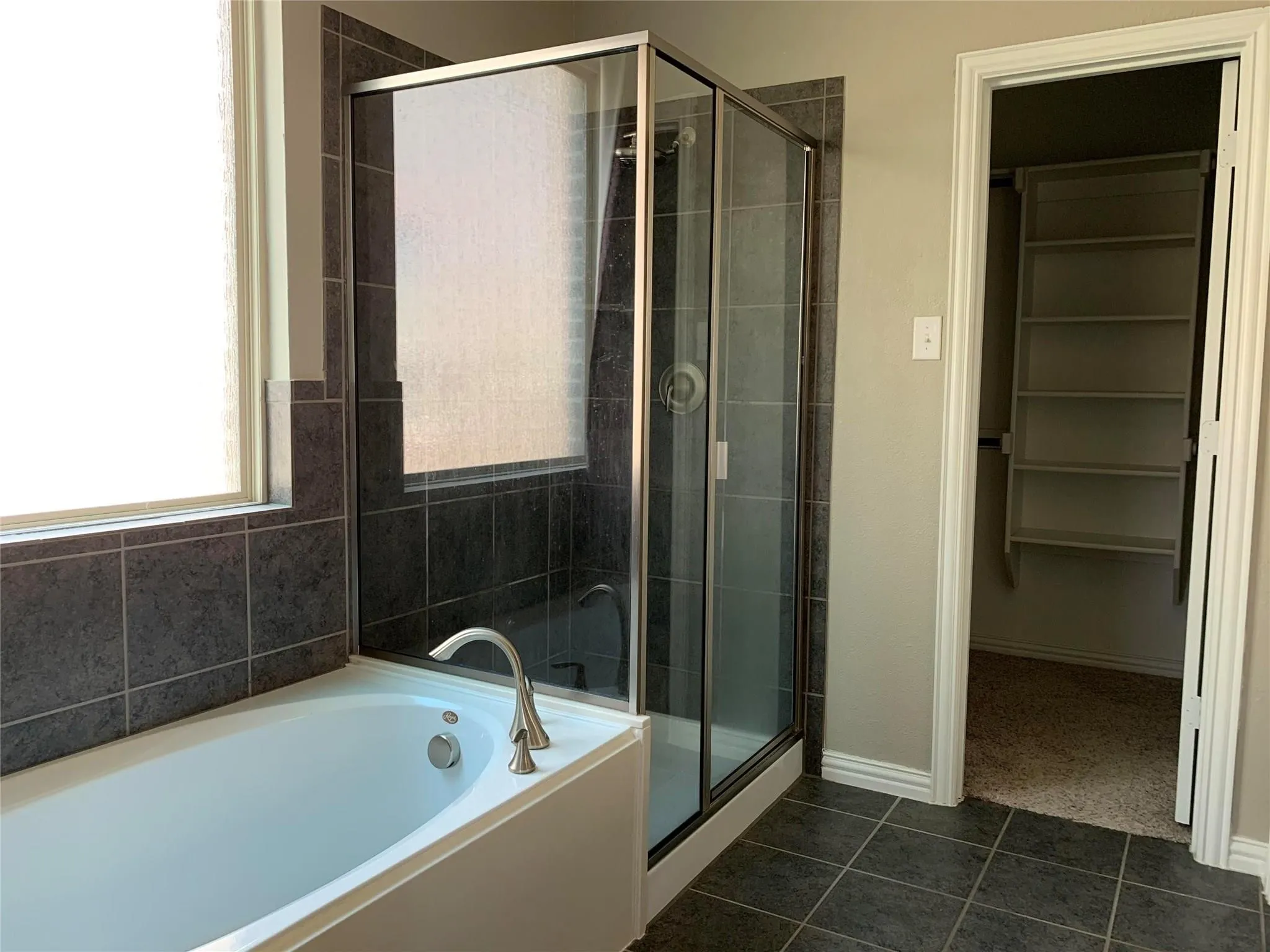 Soaking tub with brushed nickel fixtures, glass-enclosed shower with tiled surround, dark tiled flooring, and a closet with built-in shelving