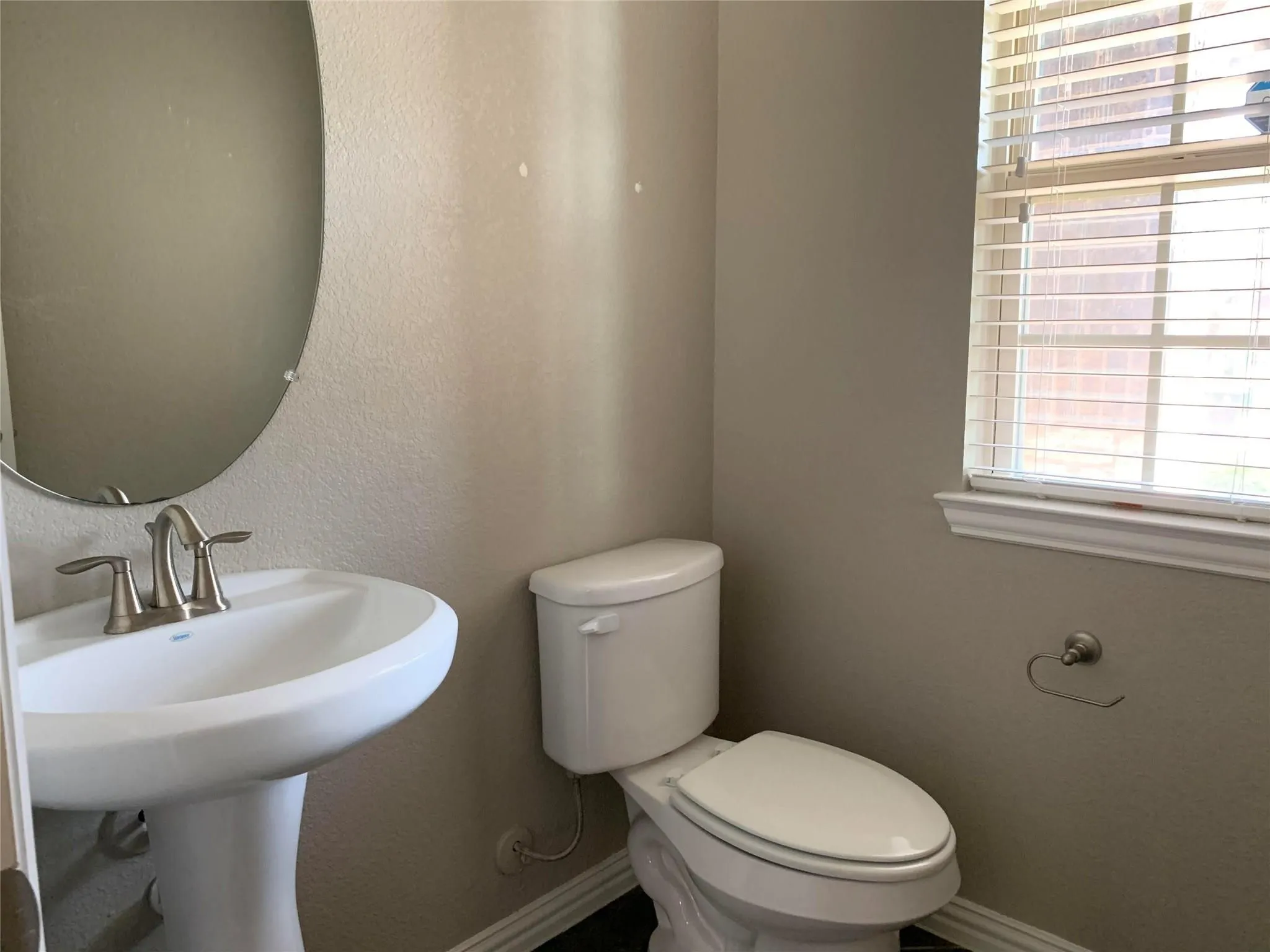 Pedestal sink with a brushed nickel faucet, window with blinds, and a circular wall mirror
