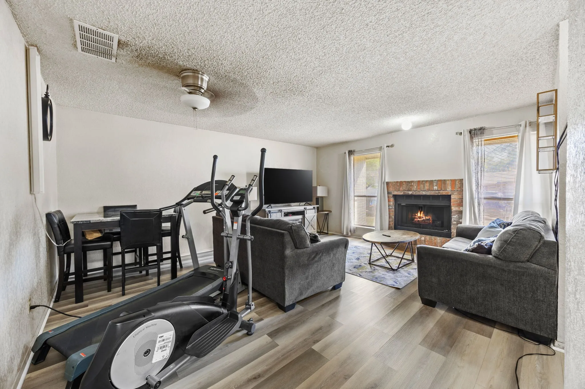 Open-concept living area with wood-finish flooring, a brick-surround fireplace, and a ceiling fan with integrated lighting