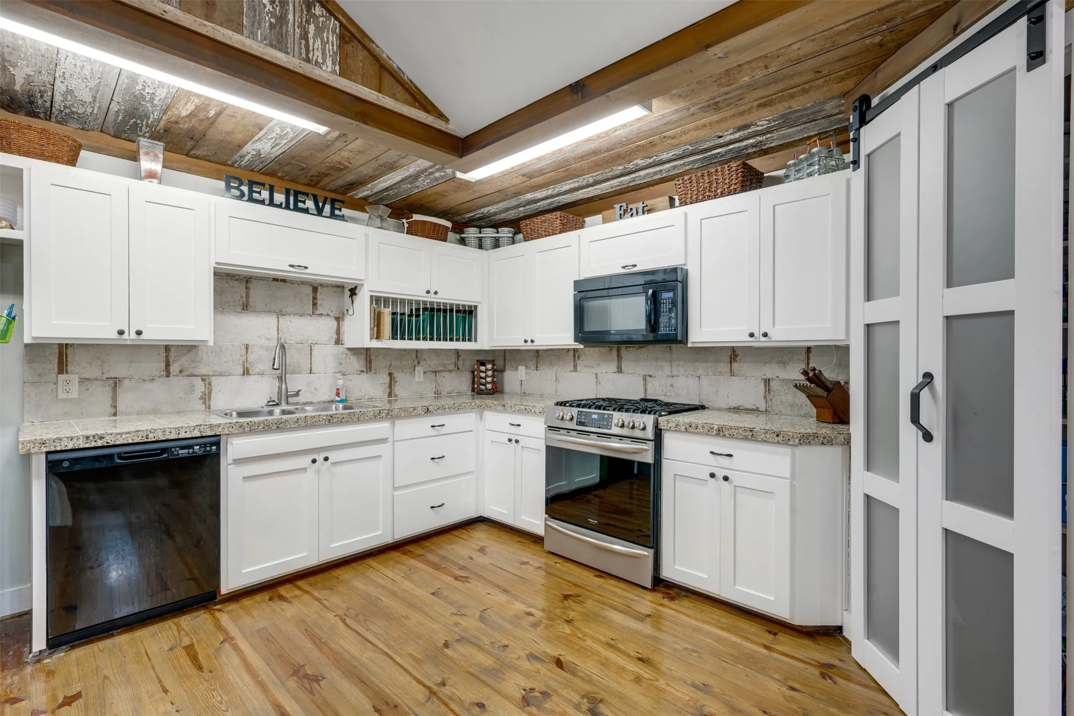 Distressed wood flooring, white cabinetry, granite-style countertops, stainless steel appliances, and a barn door with frosted glass panels