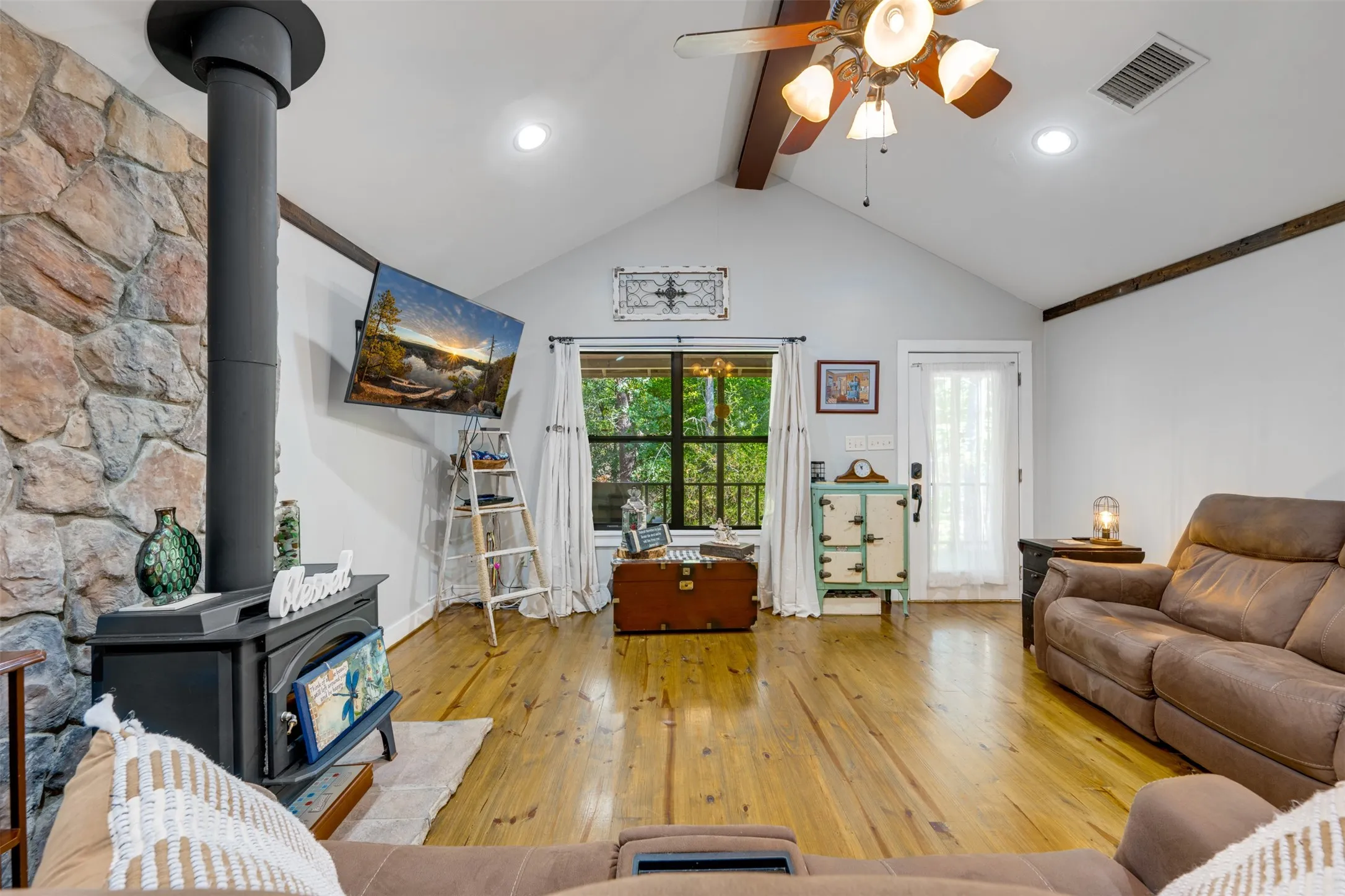 Vaulted ceiling with exposed wood beams, wood-burning stove with stone surround, wood-finish flooring, recessed lighting, and ceiling fan