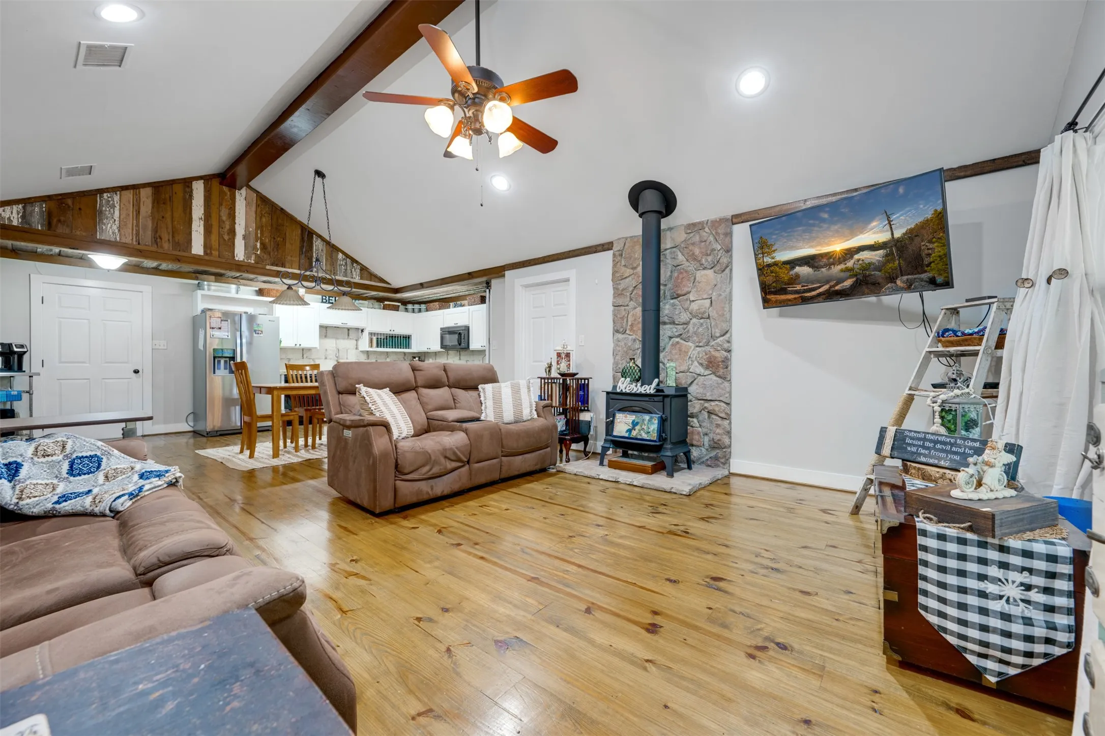 Vaulted ceiling with exposed wood beams, wood-finish flooring, stone-clad wood-burning stove, recessed lighting, and a ceiling fan