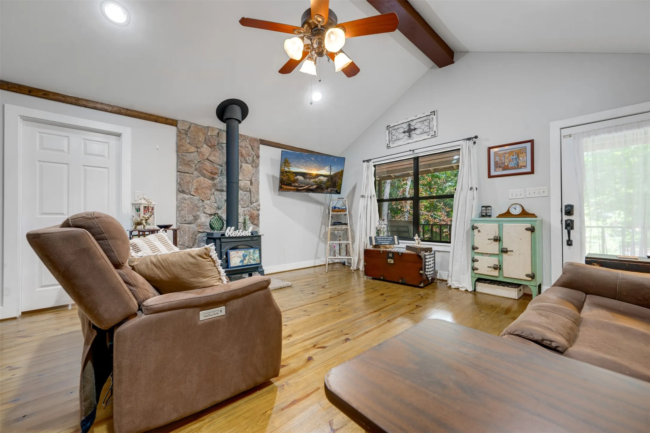 Vaulted ceiling with exposed wood beam, wood-finish flooring, stone-clad wood-burning stove, ceiling fan with integrated lighting, and multiple windows