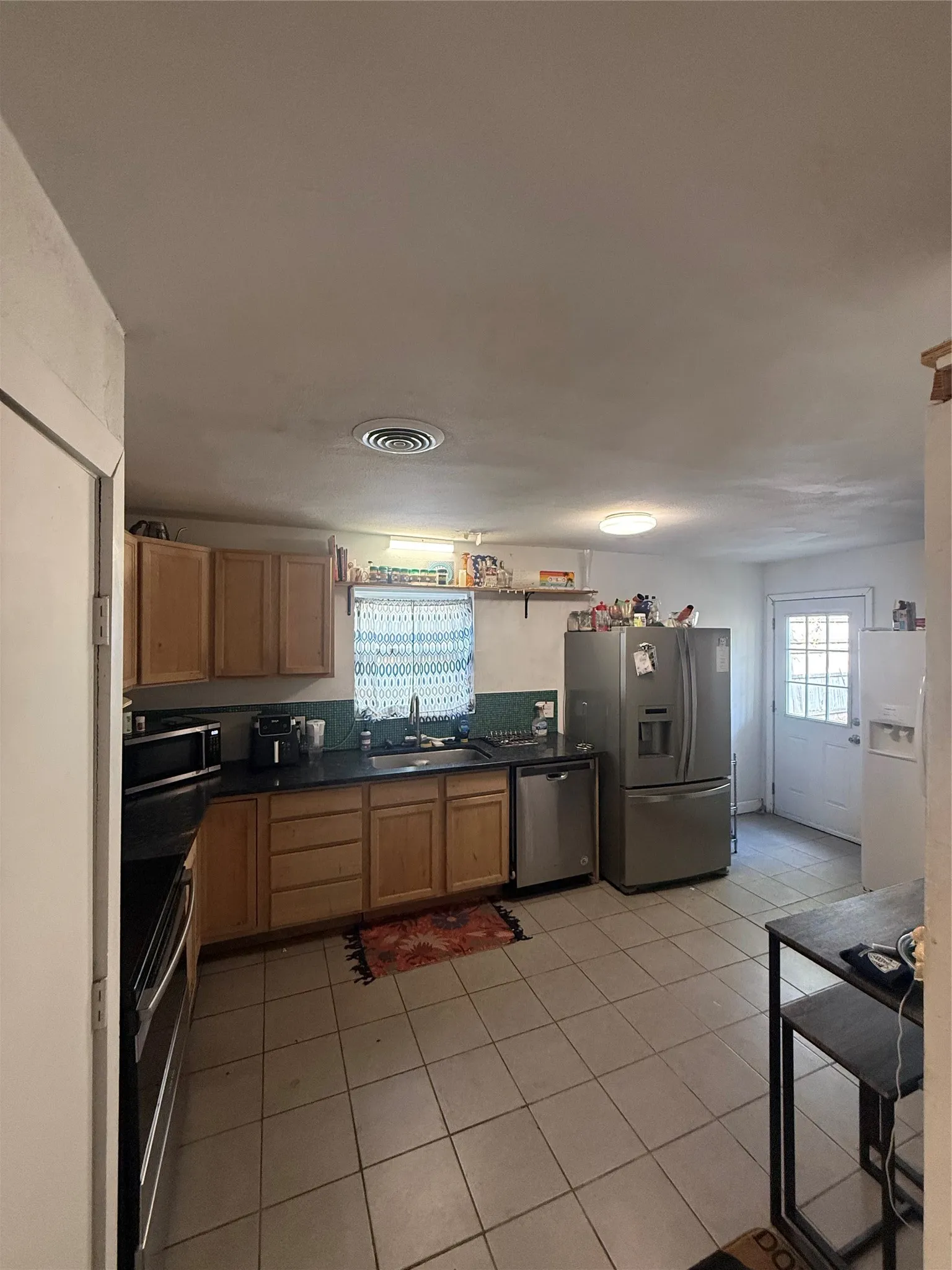 Kitchen featuring wood-finish cabinetry, black countertops, a stainless steel dishwasher, a stainless steel refrigerator, and tiled flooring