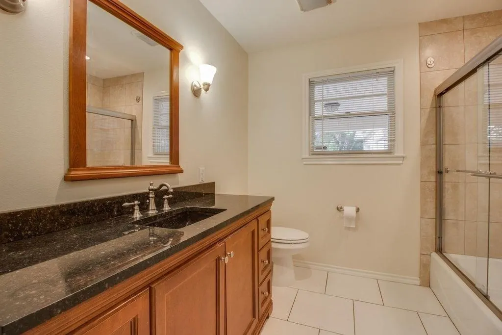 Dark-toned vanity with dual sinks and a light-toned countertop, framed mirror, wall-mounted light fixtures, tile flooring, and a tiled shower with a glass enclosure