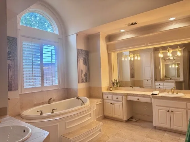 A spacious bathroom featuring a large soaking tub with integrated steps, an arched window with plantation shutters, dual vanities with integrated sinks, and ample counter space