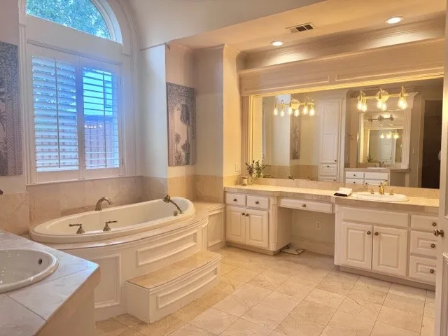 Spacious bathroom featuring a large soaking tub with steps, a vanity with dual sinks, and a window with white louvered shutters